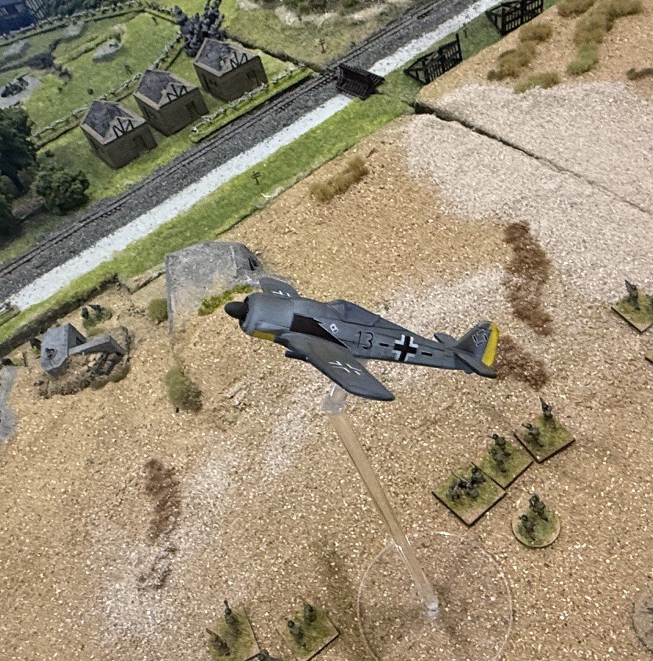 Model WWII German fighter plane flying over miniature battlefield with soldiers and terrain, set next to a model railway and small houses.