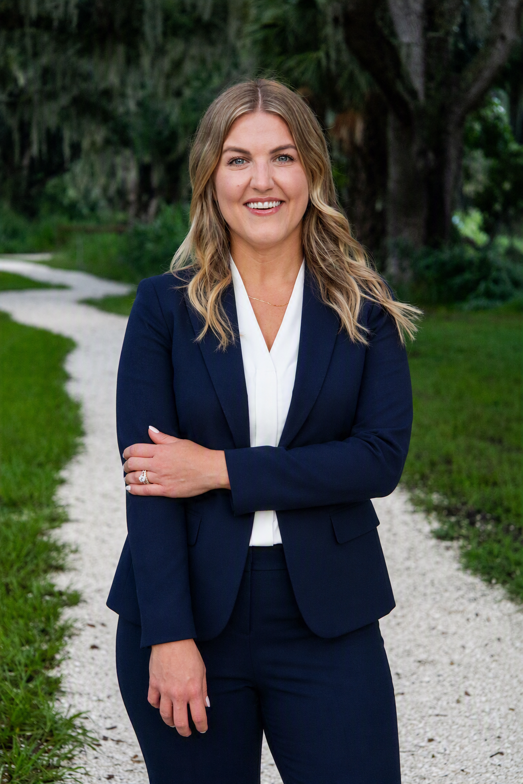 A woman in a navy blazer and white blouse standing on a gravel path in a green outdoor setting, smiling with arms crossed. Psychologist Dr. Roxanne Cole