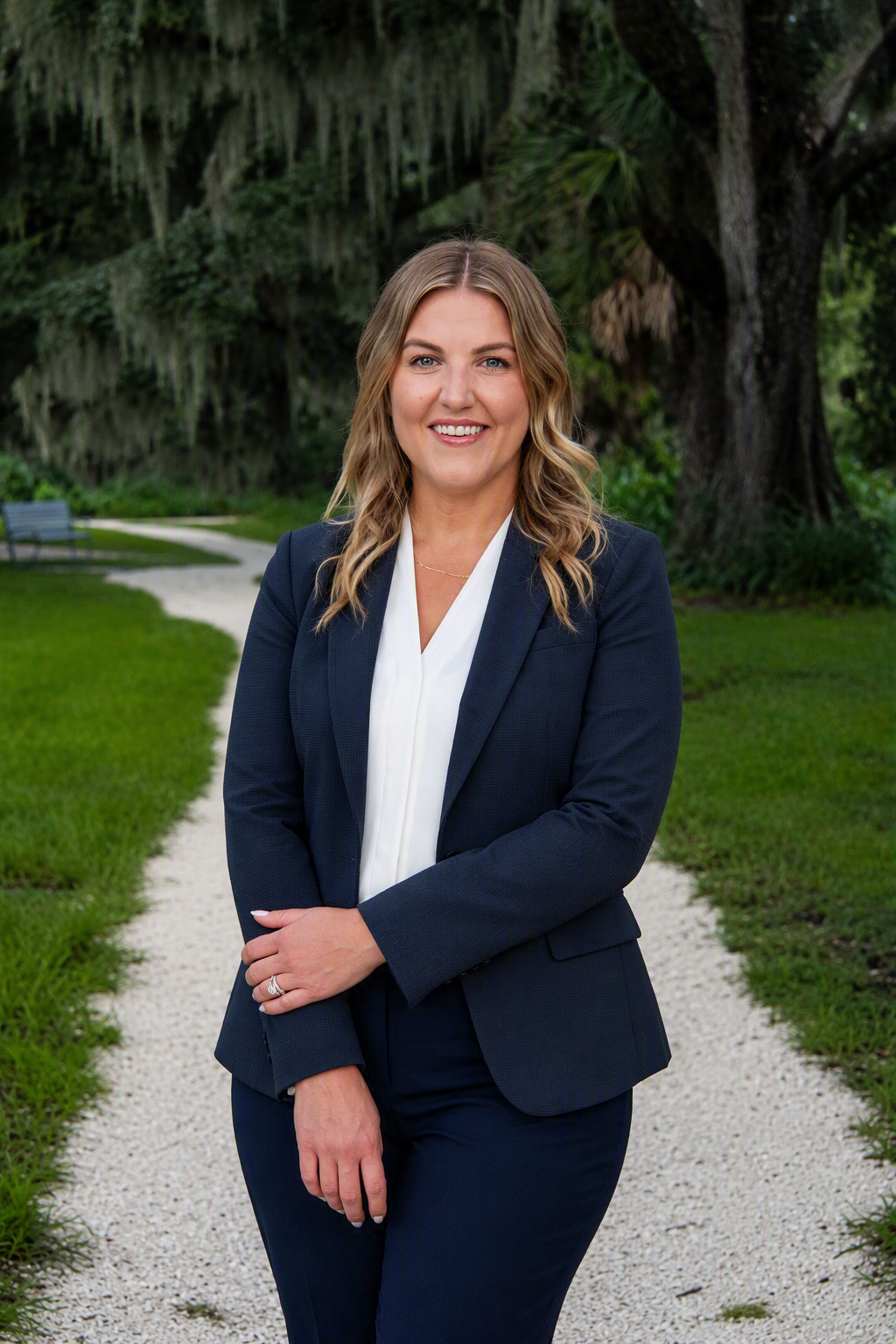 A woman with blonde, wavy hair wearing a navy blazer and white blouse standing on a gravel path in a lush, green park with trees and grass. Psychologist Dr. Roxanne Cole