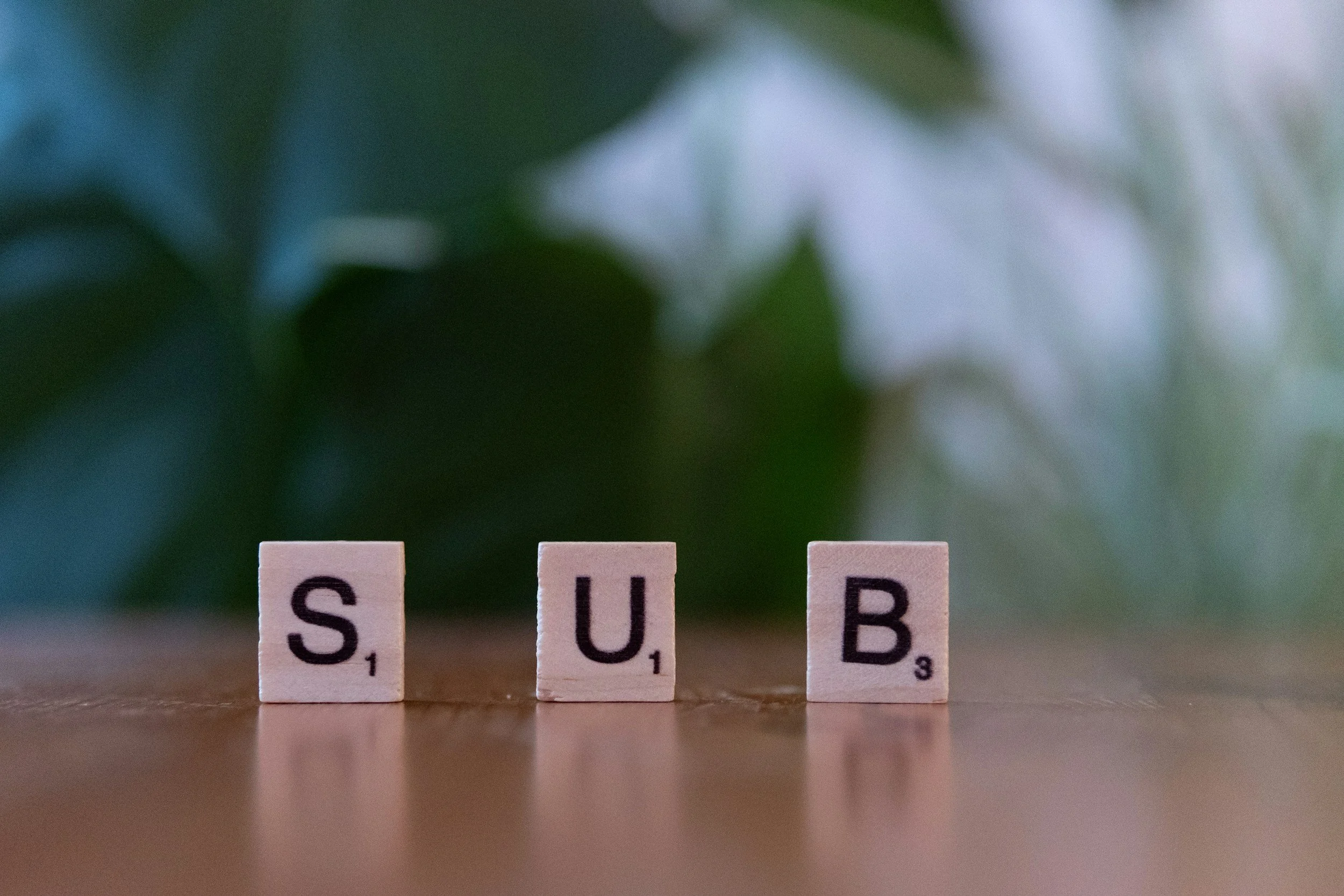 Close-up of Scrabble tiles on a wooden surface spelling out 'SUB'. In the background, blurred green leaves are visible.