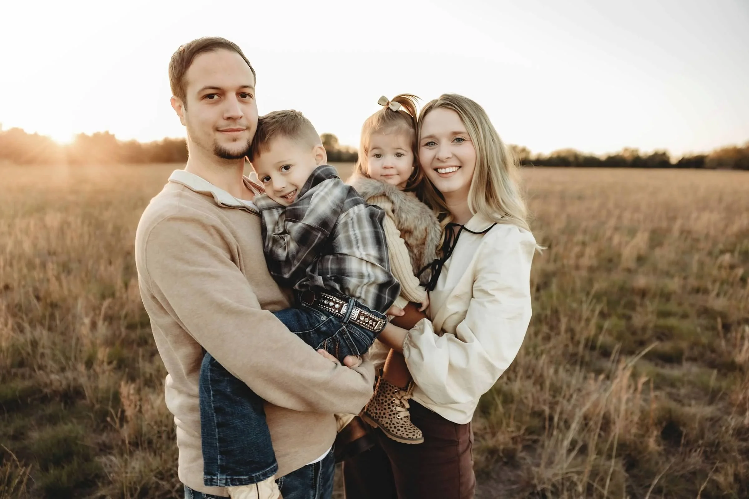 A family of four standing outdoors in a field during sunset, smiling and embracing each other. The father holds a young boy, the mother holds a young girl, and they appear happy together.