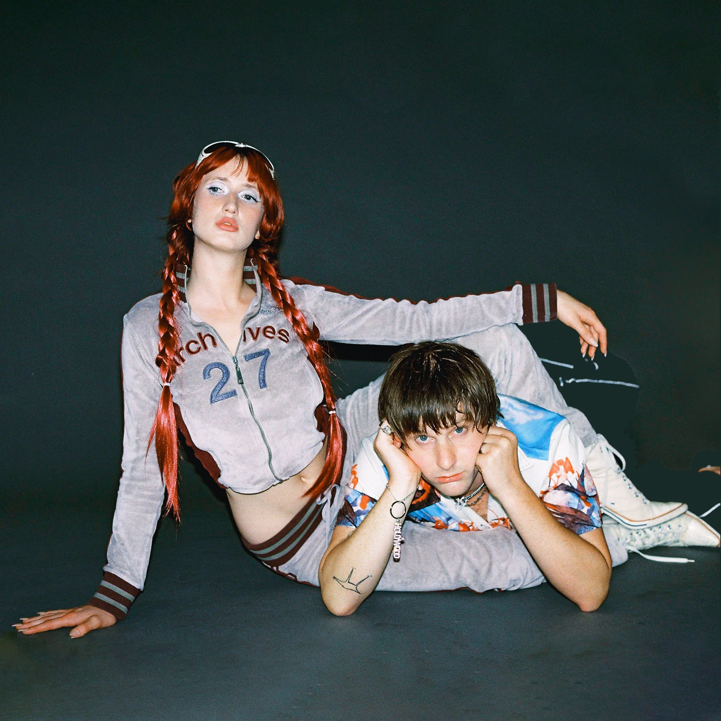 A young woman with red hair styled in braids with a headphone on her head, wearing a zip-up jacket with text on it, and a young man with brown hair and tattoos on his arm, wearing a colorful shirt, lying on a dark surface.