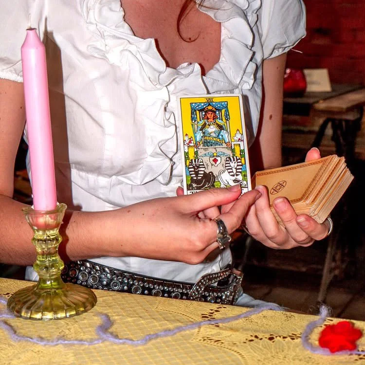 A woman wearing a white ruffled blouse holds a Tarot card deck and a single tarot card featuring The Chariot card, with a pink candle in a gold candle holder on a table covered with a yellow cloth with purple and red accents.