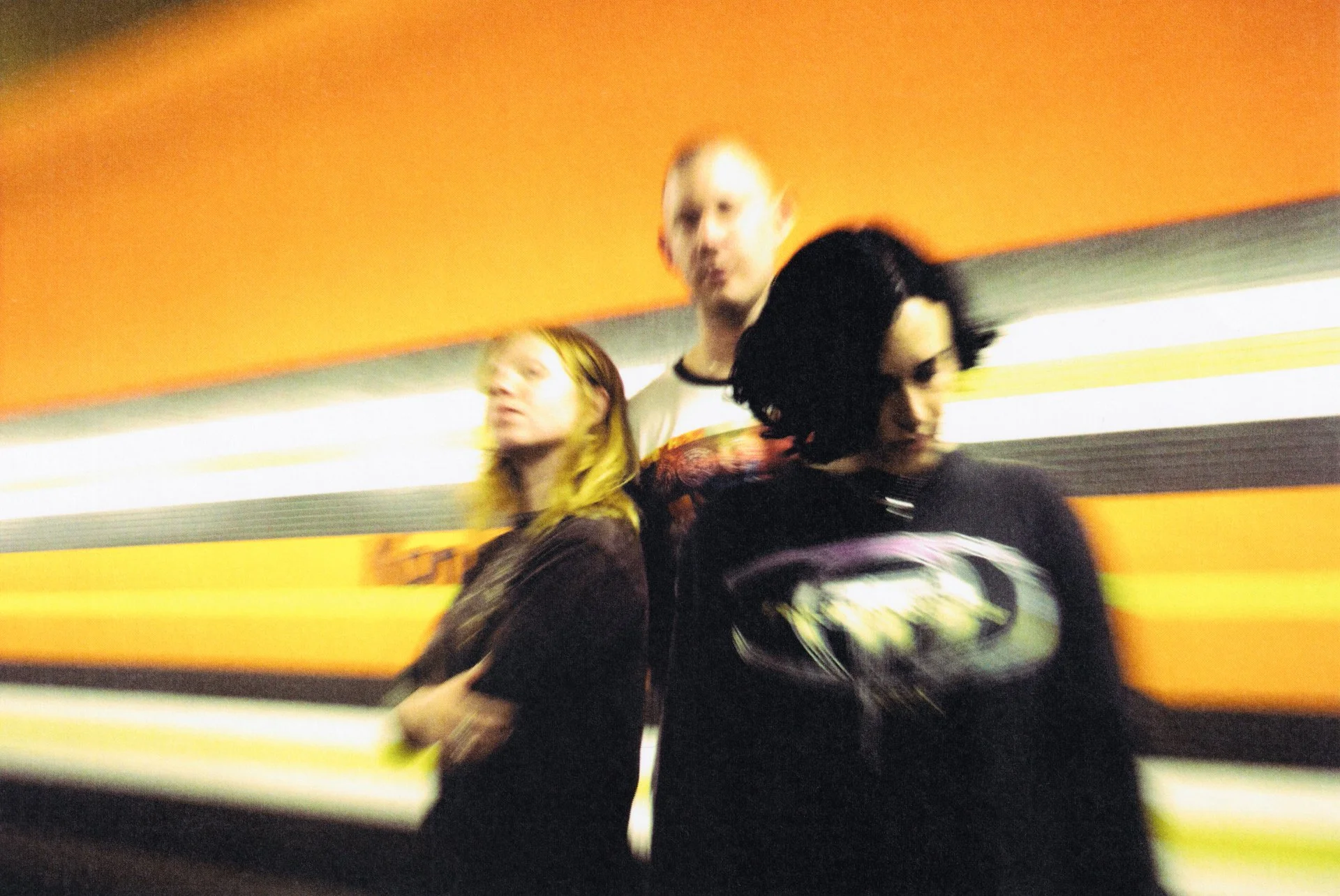 Three people walking in an UBahn station with an orange wall and illuminated background bands. The photo is blurry and motioned.