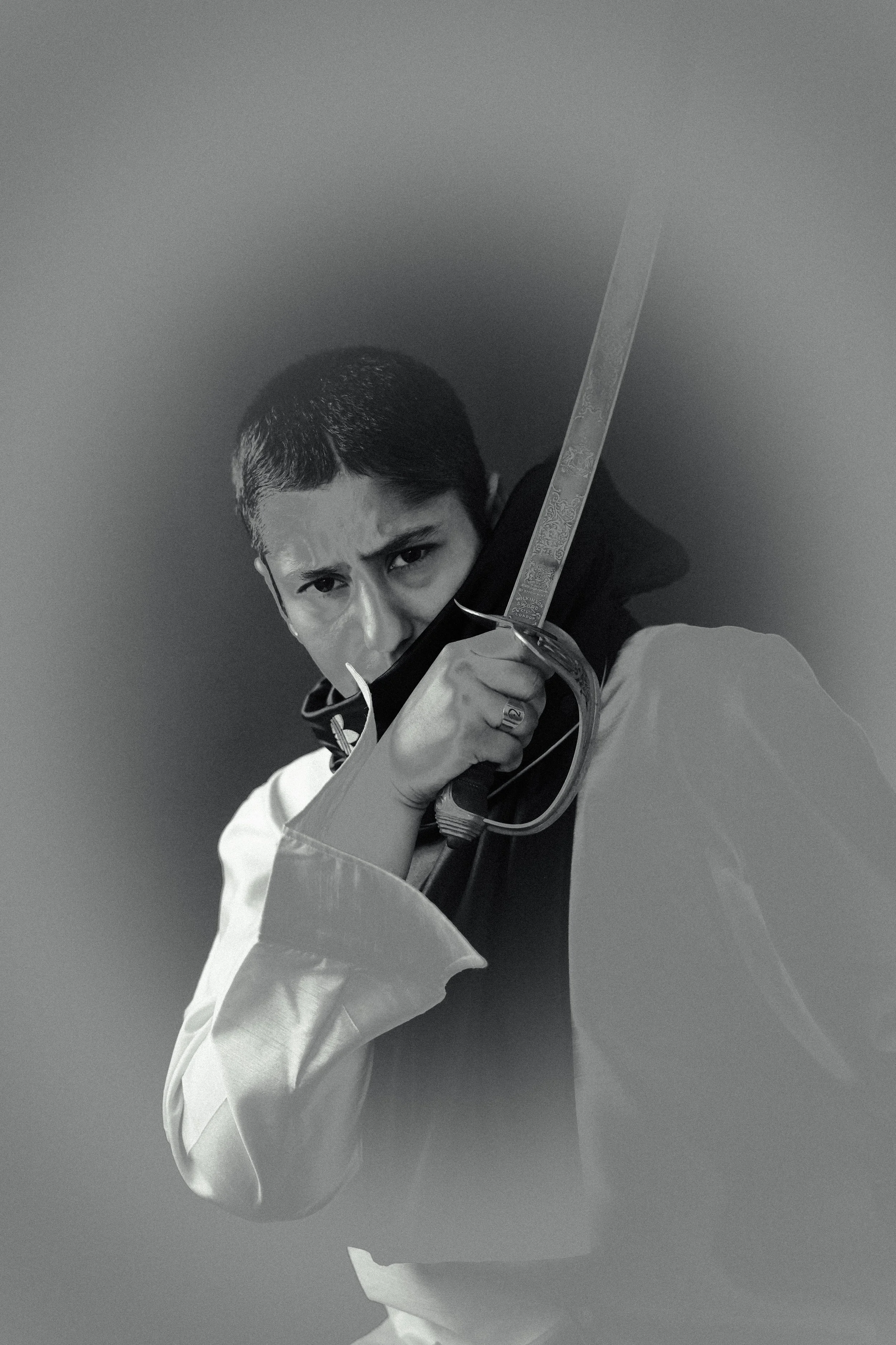 A person with short hair holding a sword with both hands, aiming it forward in a serious pose, wearing a white robe with a black collar.