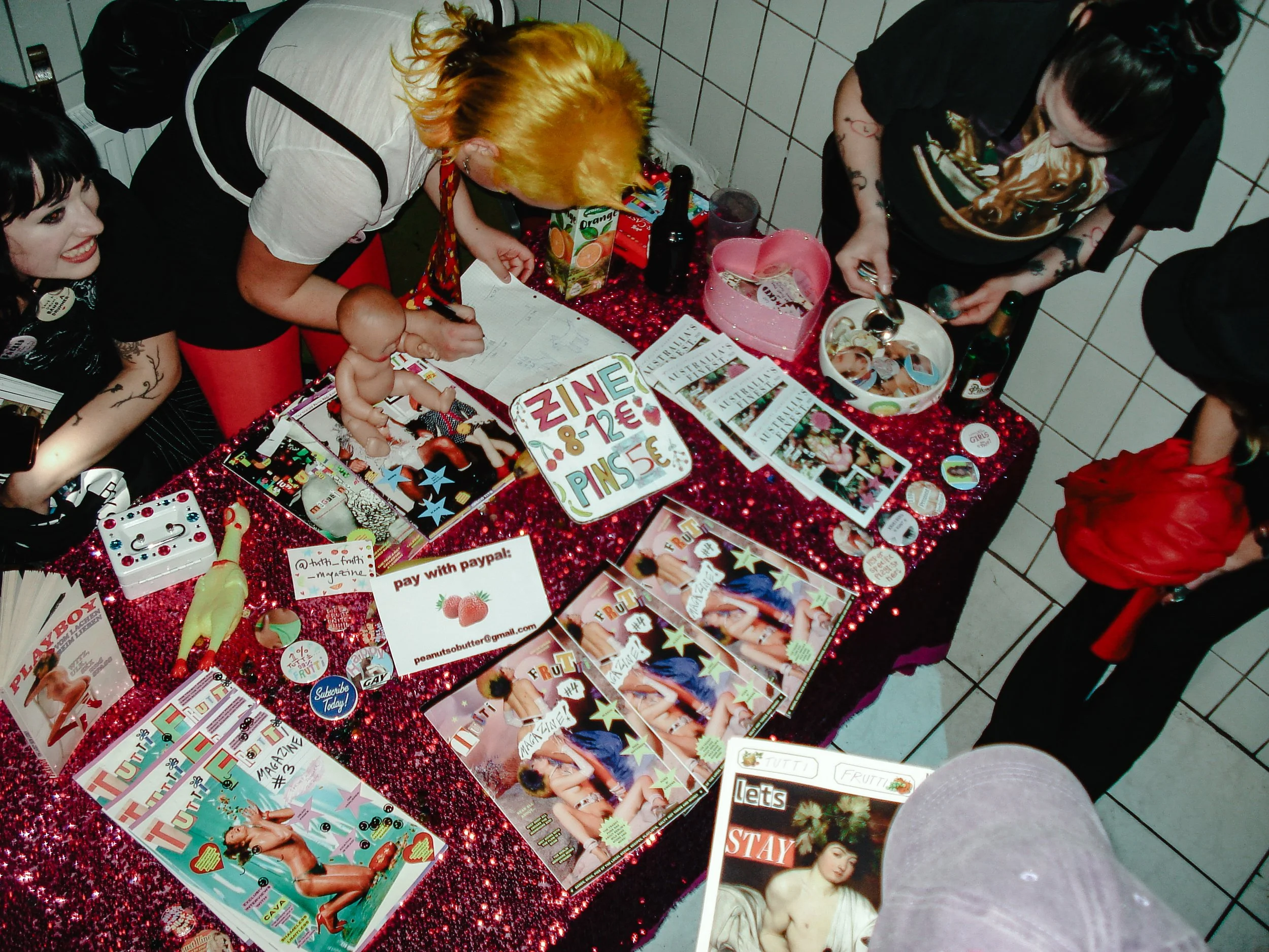 People gather around a table decorated with a red sparkly tablecloth, magazines, and various small items, at a party or event.