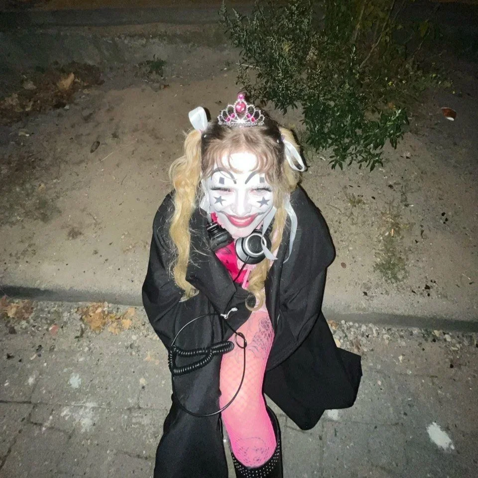 Full body photograph of someone sitting on a curb wearing a pink tiara and with black & white geometric face paint.