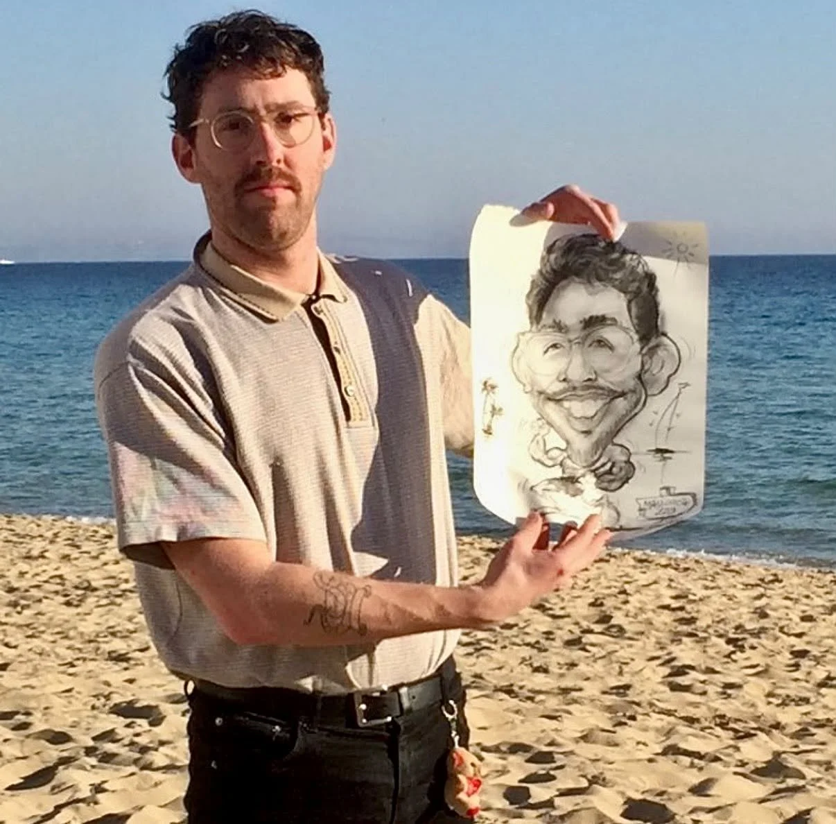 A man with glasses and a beard holding a caricature drawing of himself on a beach with the ocean in the background.