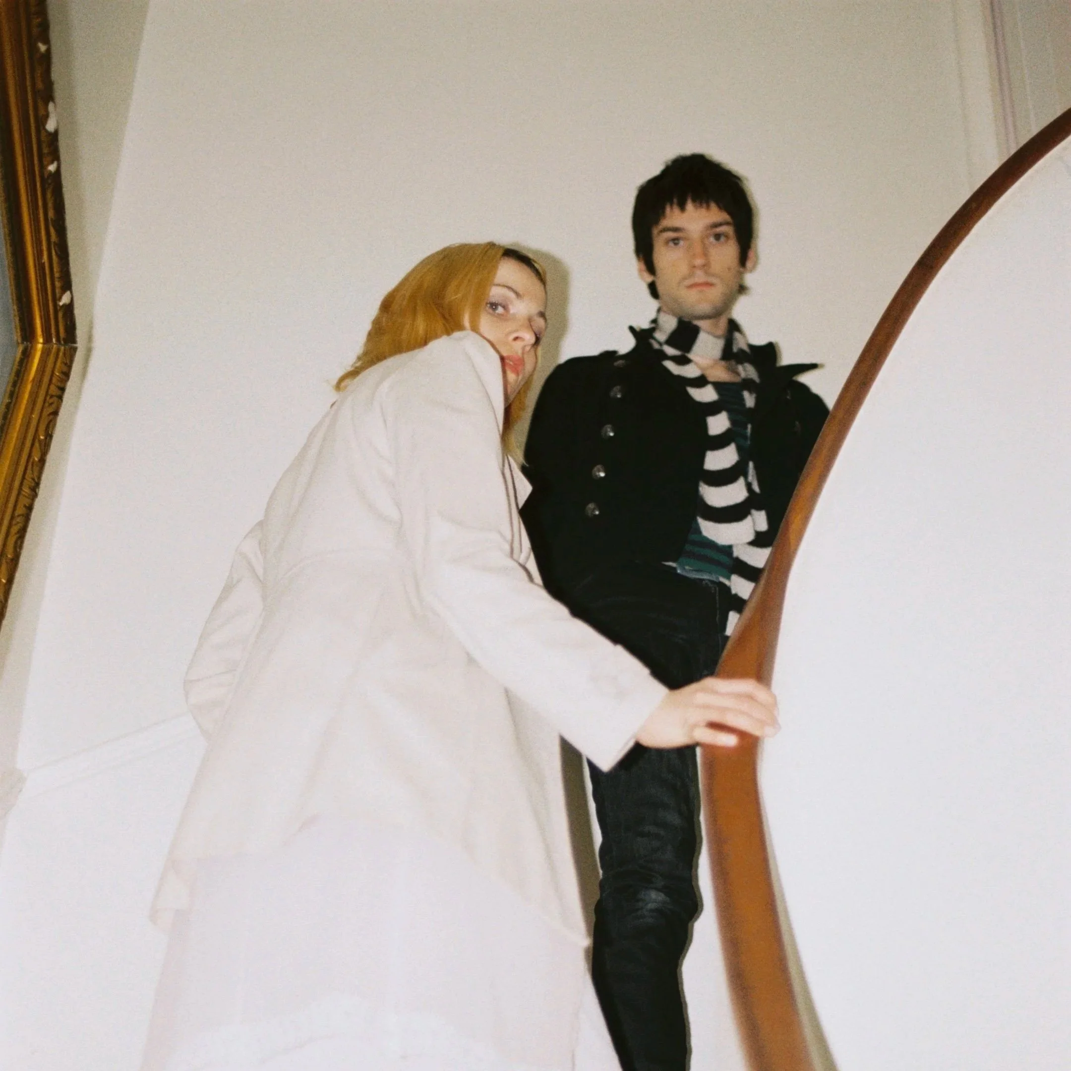 A young woman with red hair and a man with dark hair and a striped shirt stand on a staircase with a wooden banister, looking at the camera with serious expressions.