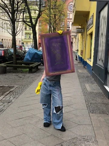 A full body photograph of a person wearing ripped jeans & rubber gloves holding up a dirtied screen printing screen over their face & upper body.
