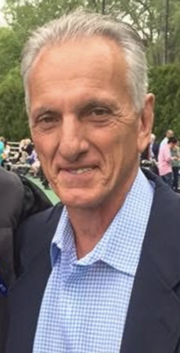 A smiling middle-aged man with gray hair in a suit and light blue collared shirt outdoors, with blurred trees and people in the background.