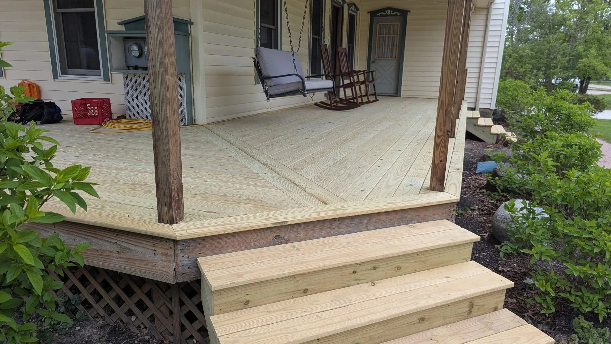 A newly built wooden front porch with steps, railing posts, a swing, and rocking chairs, surrounded by green bushes and plants.