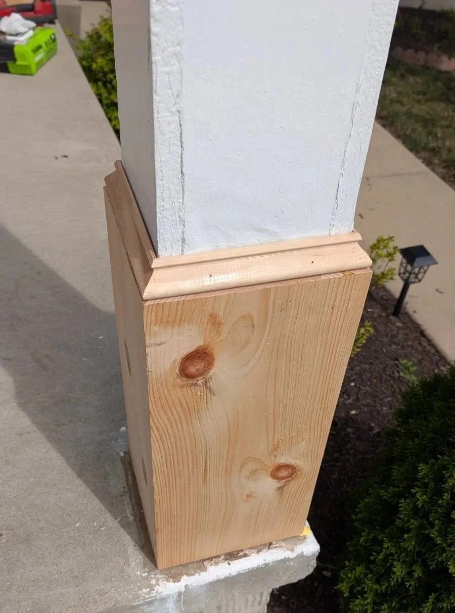 Close-up of a wooden post with a decorative trim at the top, painted white on the upper section, located outdoors on a concrete surface near some plants and a lamp post.