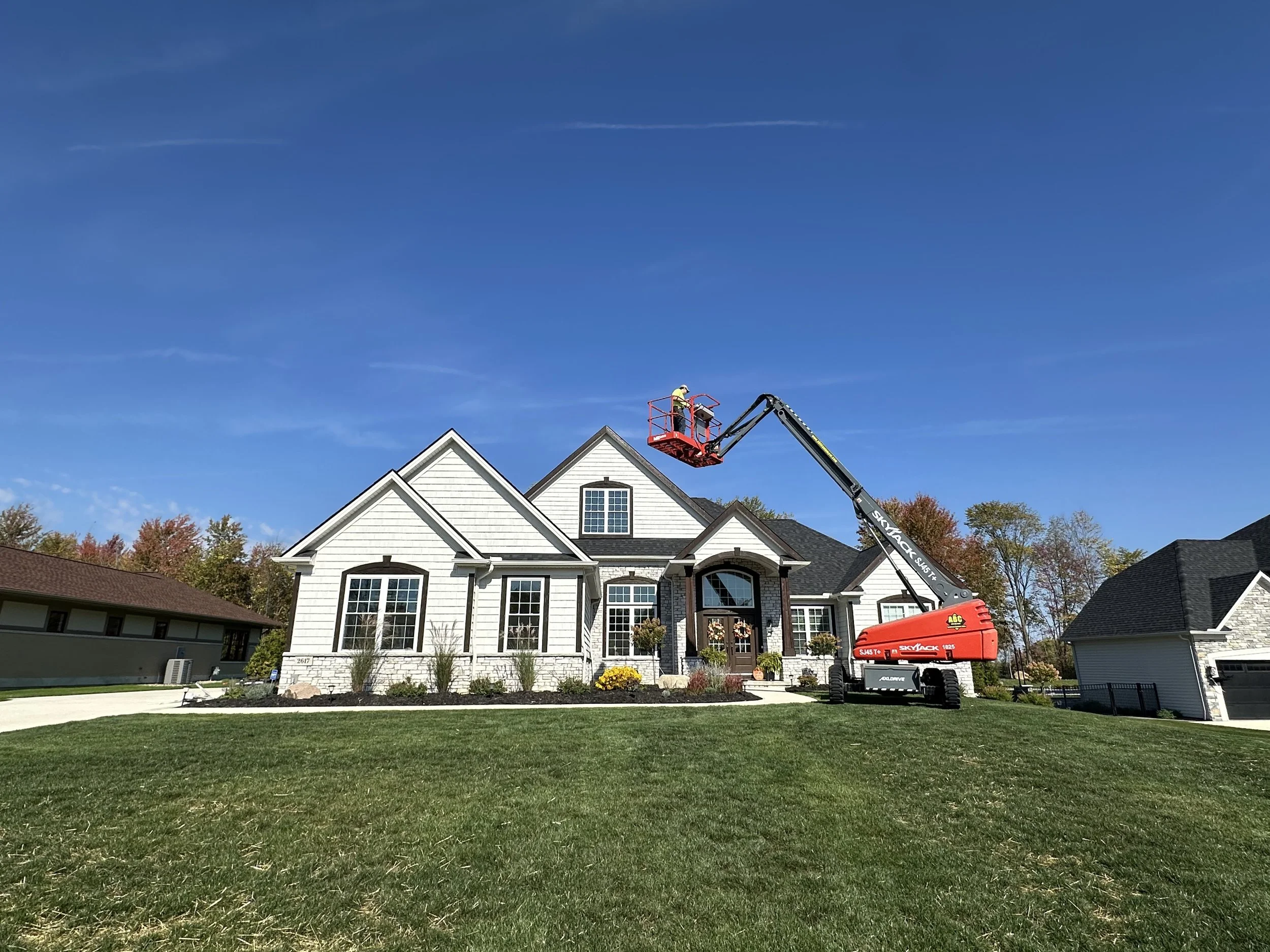 A house with a white exterior and multiple gabled roofs is being worked on with a lift. A worker is in the lift working on the upper part of the house under a blue sky.