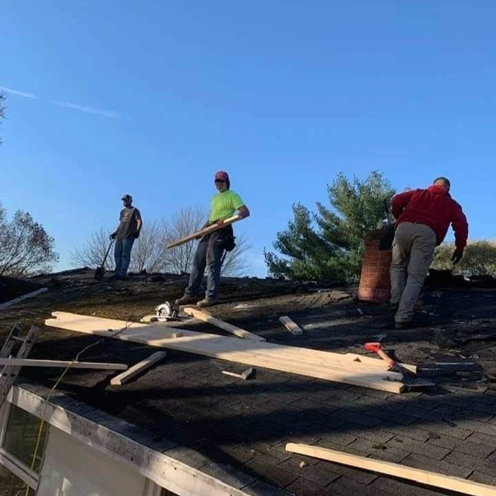 Three construction workers repairing a roof. One stands holding a wooden plank, another is on the roof near a chimney, and the third is in the background near the roof's edge.