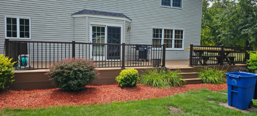 Back porch with black railings, wooden steps, white house exterior, green shrubs, mulch, grass, blue recycling bin, and garden hose.