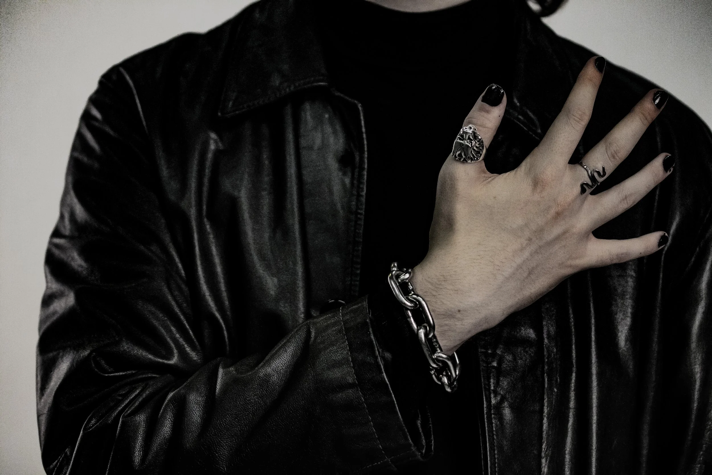Close-up of a person's hand wearing rings and a bracelet, with black nail polish, resting on a black leather jacket.