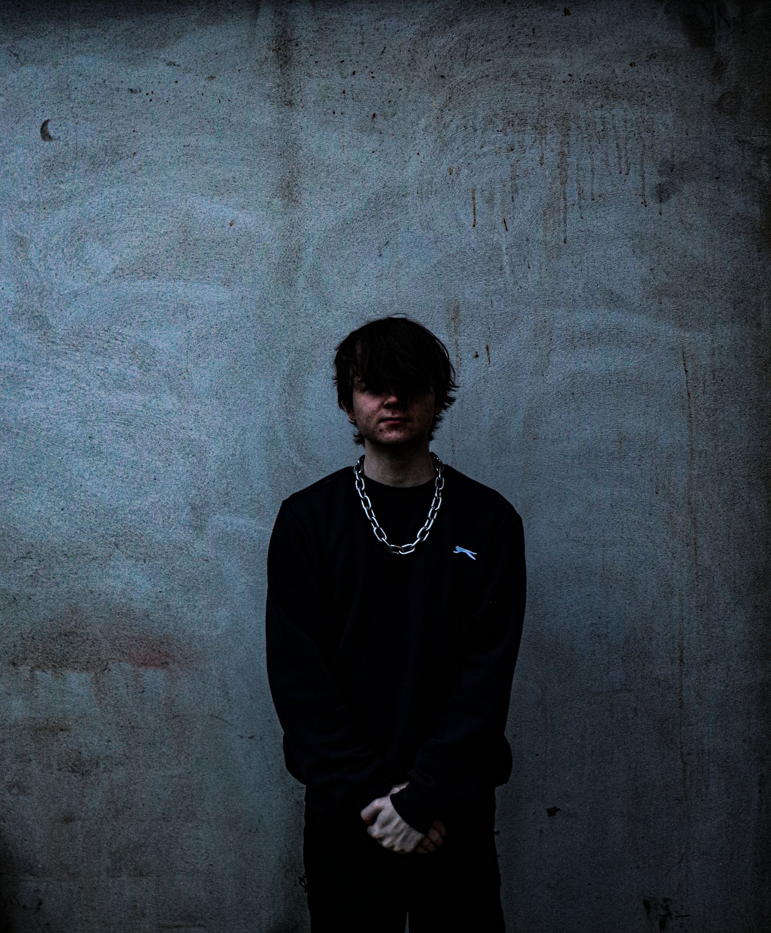 A young person with dark, messy hair, wearing a black shirt and a silver chain necklace, stands against a textured concrete wall. The individual has a neutral expression, their arms crossed, and looks slightly downward.