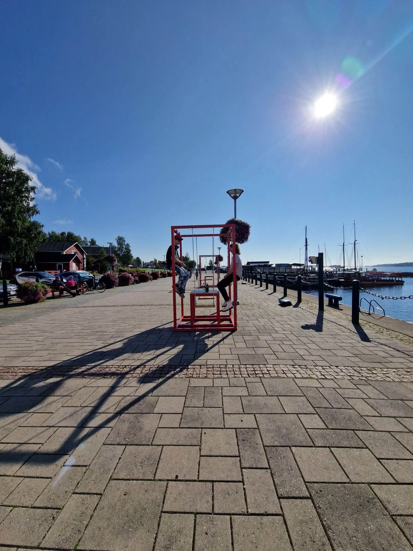 Two children are playing on a red swing set near a waterfront, with boats docked at a marina, under a clear blue sky with the bright sun shining.