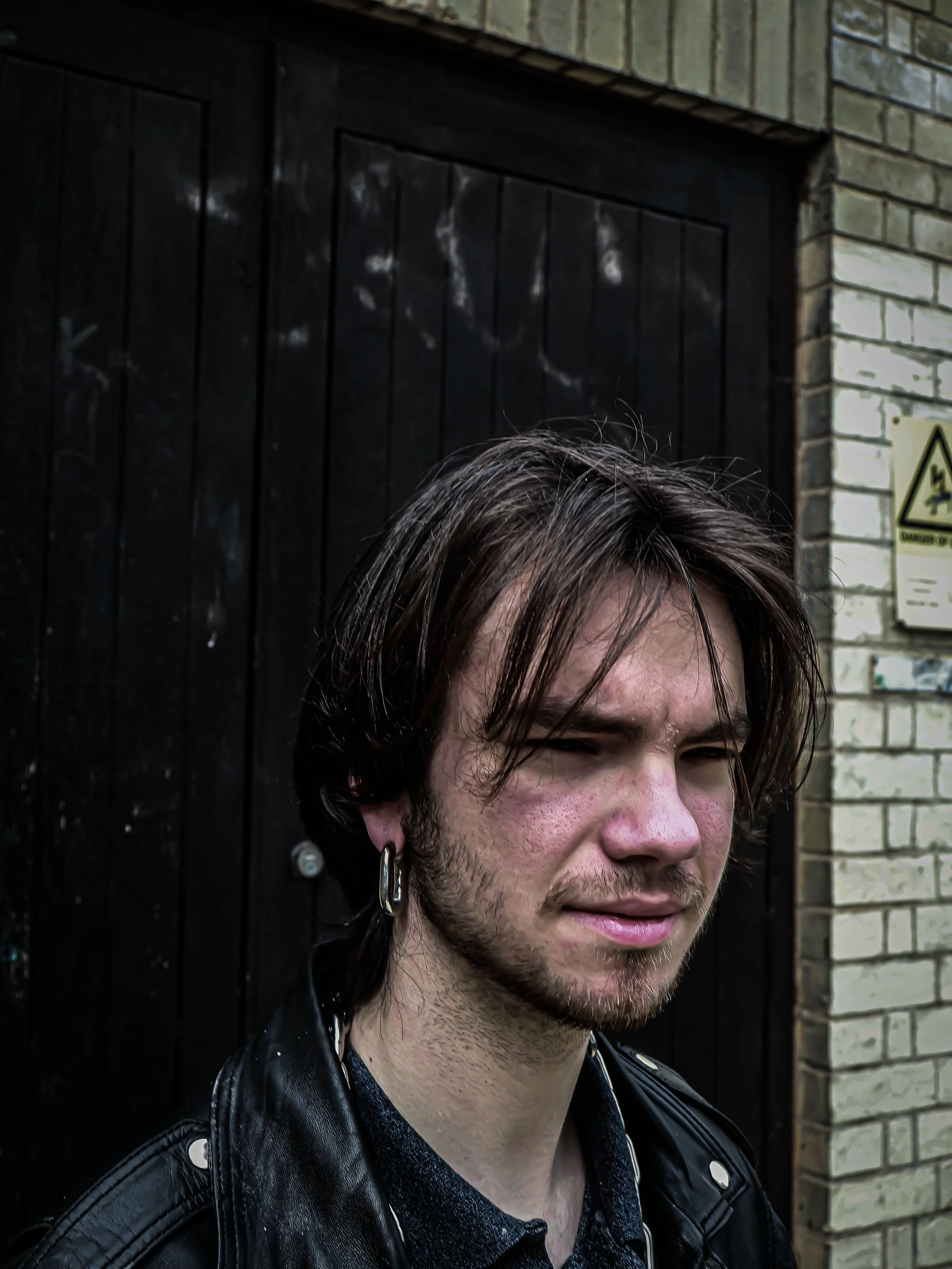 A young man with dark brown hair, facial hair, and earrings, standing outdoors in front of a brick wall and a black door with graffiti.
