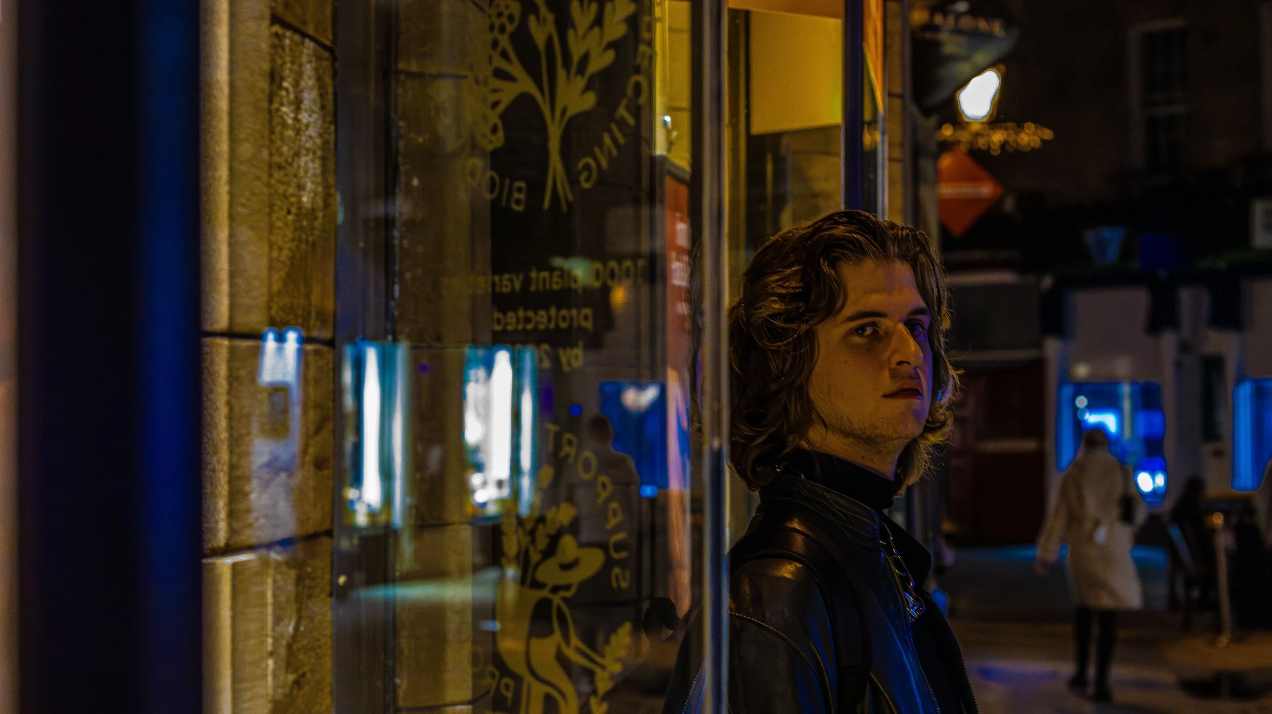 A young man with long, wavy hair and a leather jacket standing outside at night and looking at the camera, with a storefront window with yellow text and a yellow logo reflecting on it nearby.