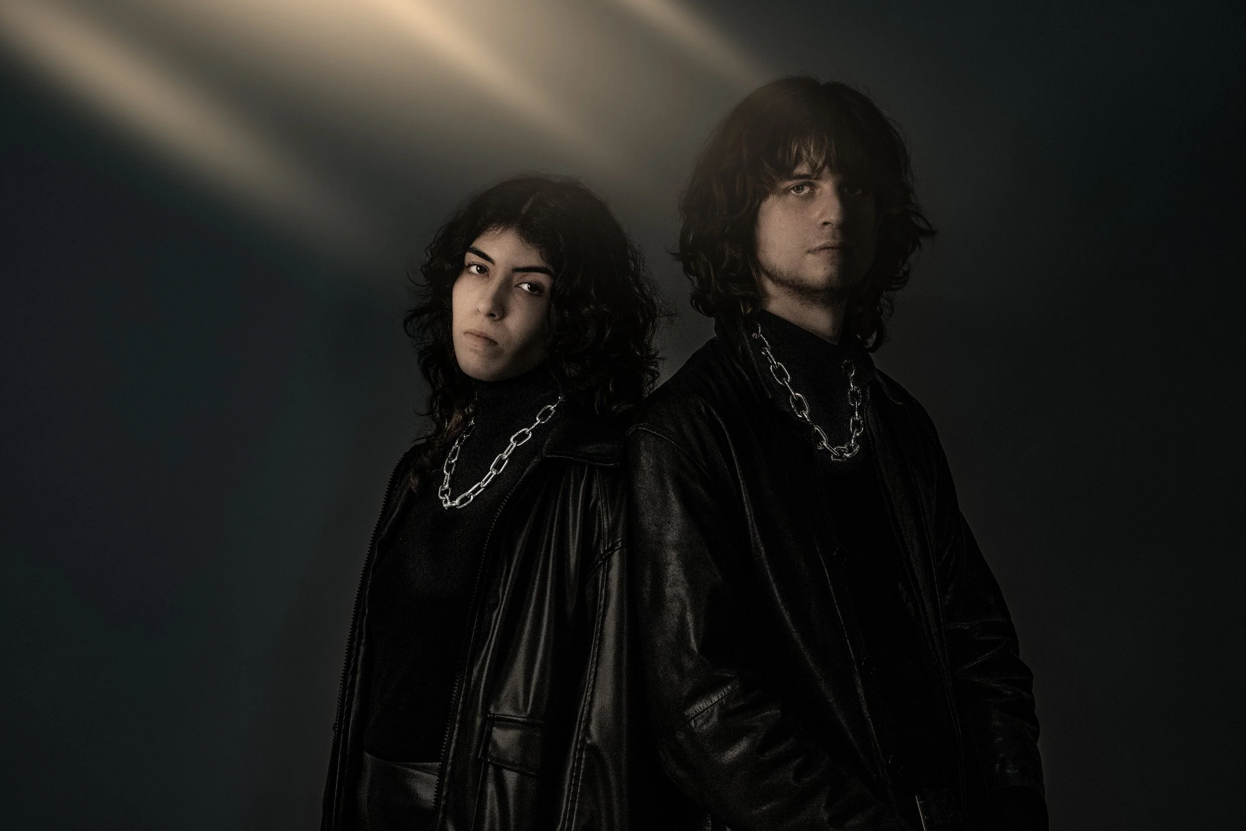 Two young adults, one male and one female, with dark, wavy hair, wearing black leather jackets and silver chains, standing against a dark background with dramatic lighting.
