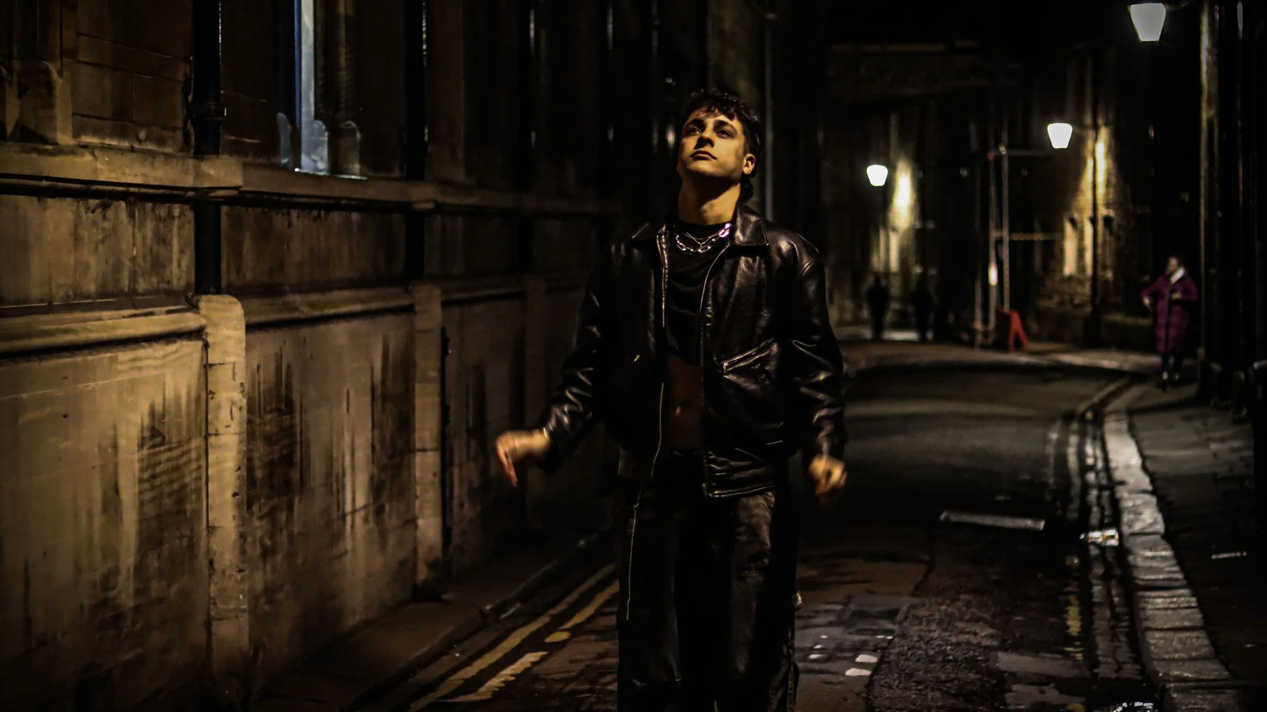 A young man walking down a dark, wet city street at night, wearing a black leather jacket and layered necklaces, with streetlights illuminating the scene.