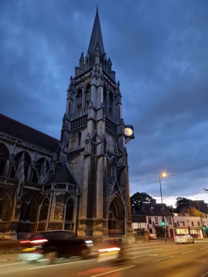A tall, Gothic-style church with a clock tower and spire, set against a dark blue evening sky, with cars passing by on the street in the foreground.