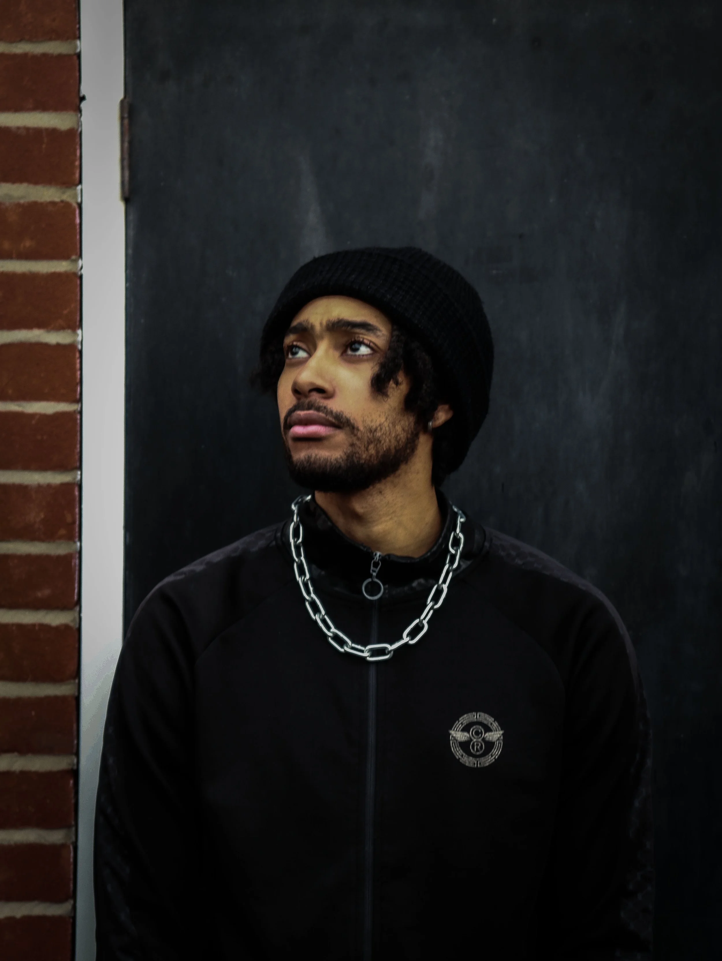 A man with curly hair and a mustache wearing a black beanie, a chunky chain necklace, and a black jacket with a circular logo on the chest, standing against a black background with a brick wall on the left.