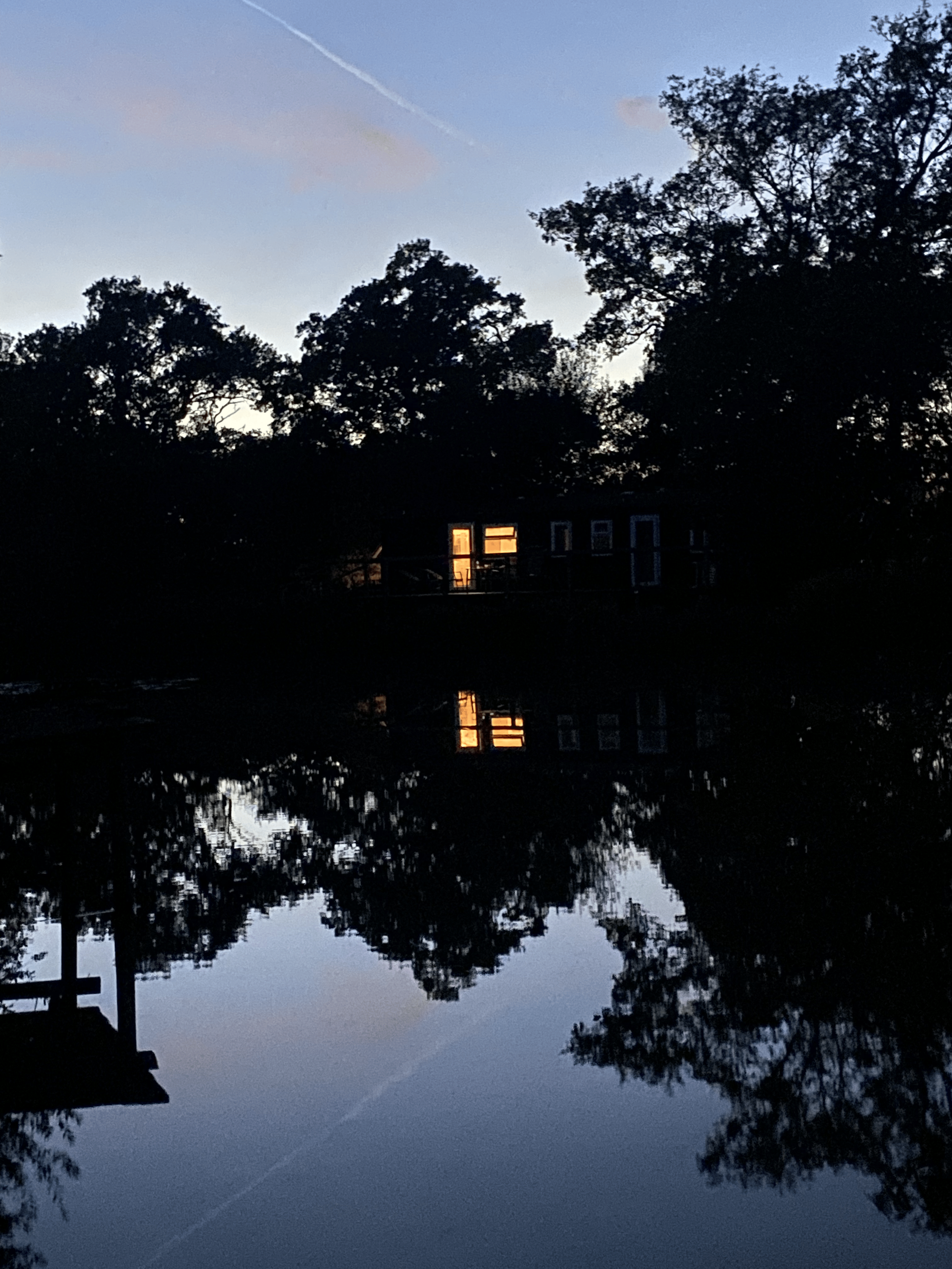 Sunset scene with trees and a house reflected in a calm body of water; sky showing light blue and gray hues with wispy clouds and a contrail.