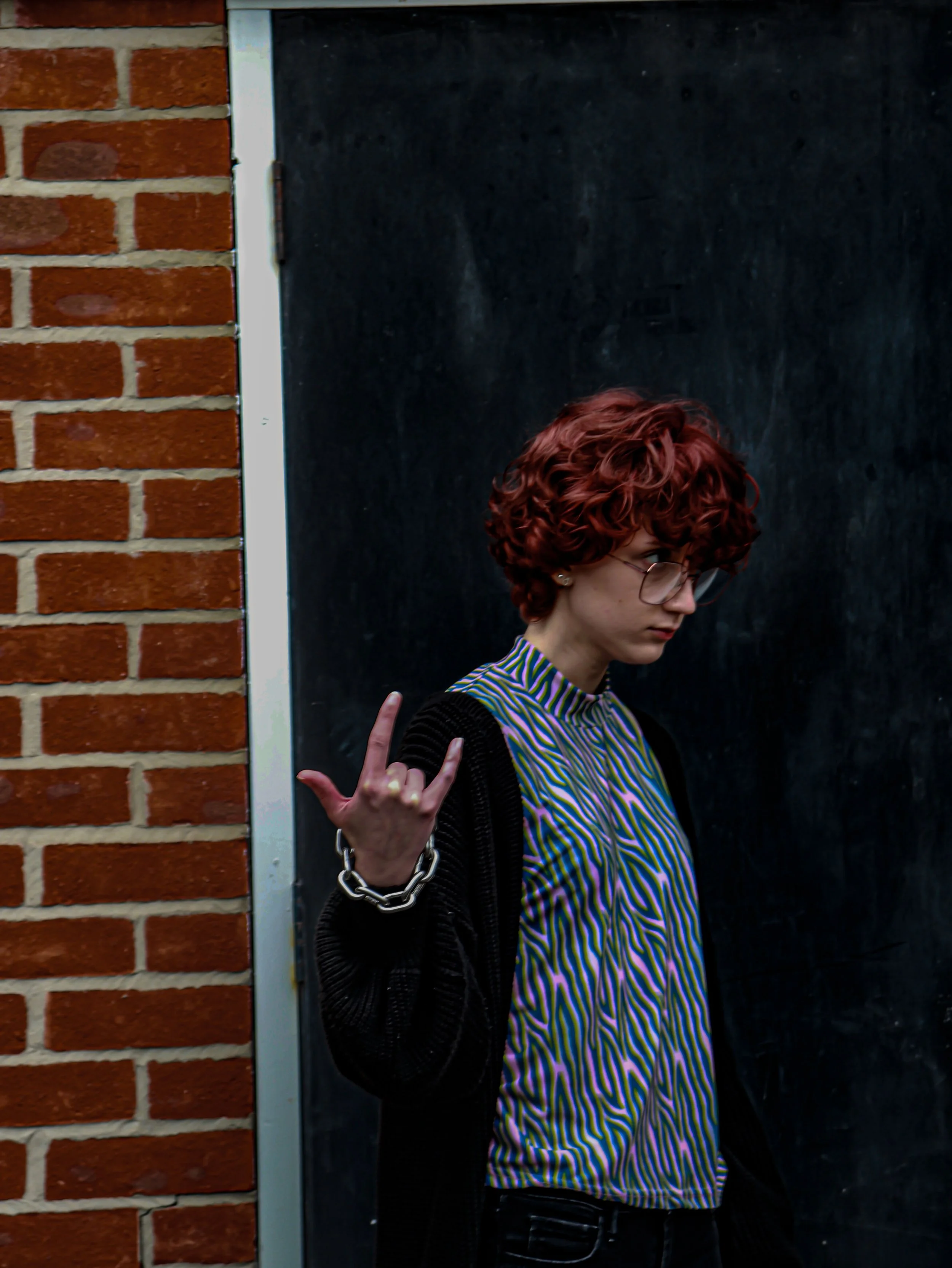 A woman with curly red hair and glasses standing against a black surface and a brick wall, making a peace sign with her right hand.