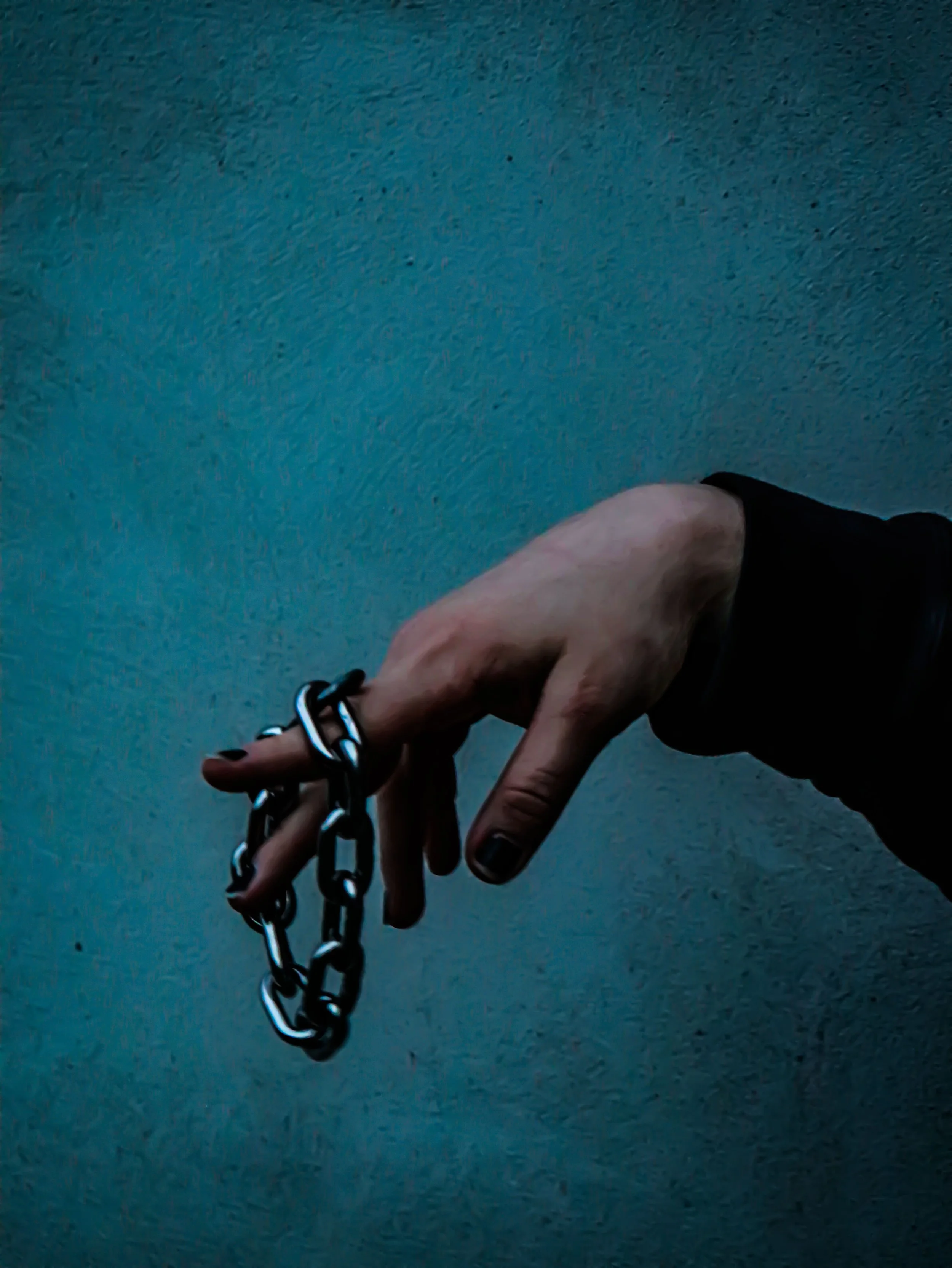 A hand holding a chain in front of a teal textured wall, with part of a person's arm visible in dark clothing.