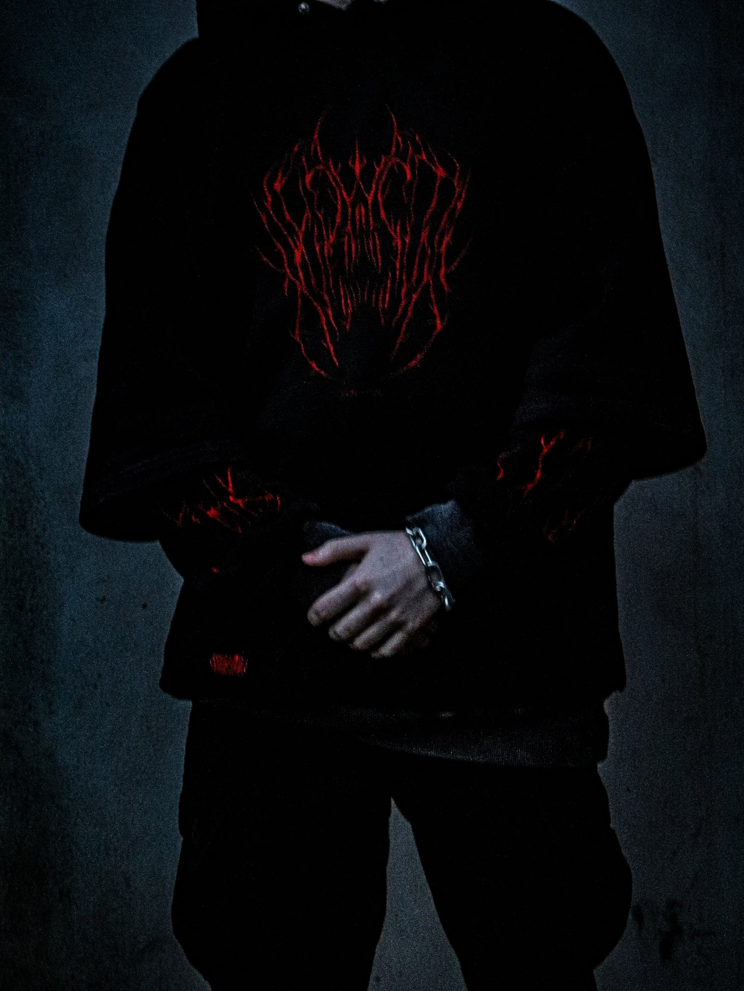 Person wearing a black hoodie with red intricate design on the chest, in a dark setting, with the hand of the person touching the fabric.