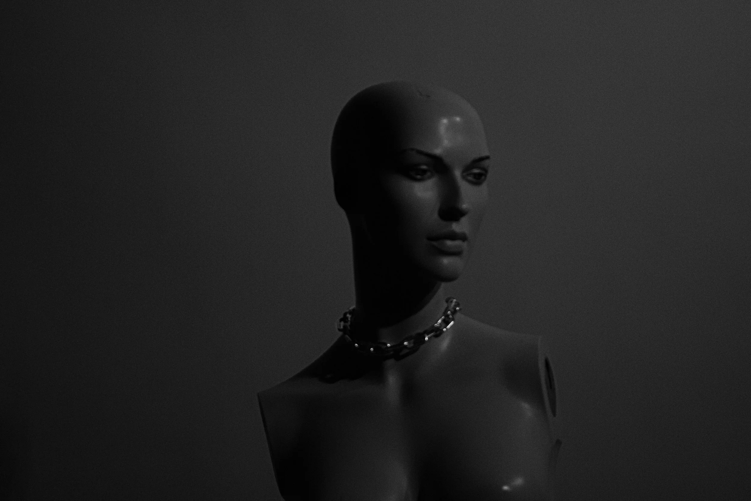 Black mannequin head with short hair wearing a chunky chain necklace, set against a dark background with soft lighting.