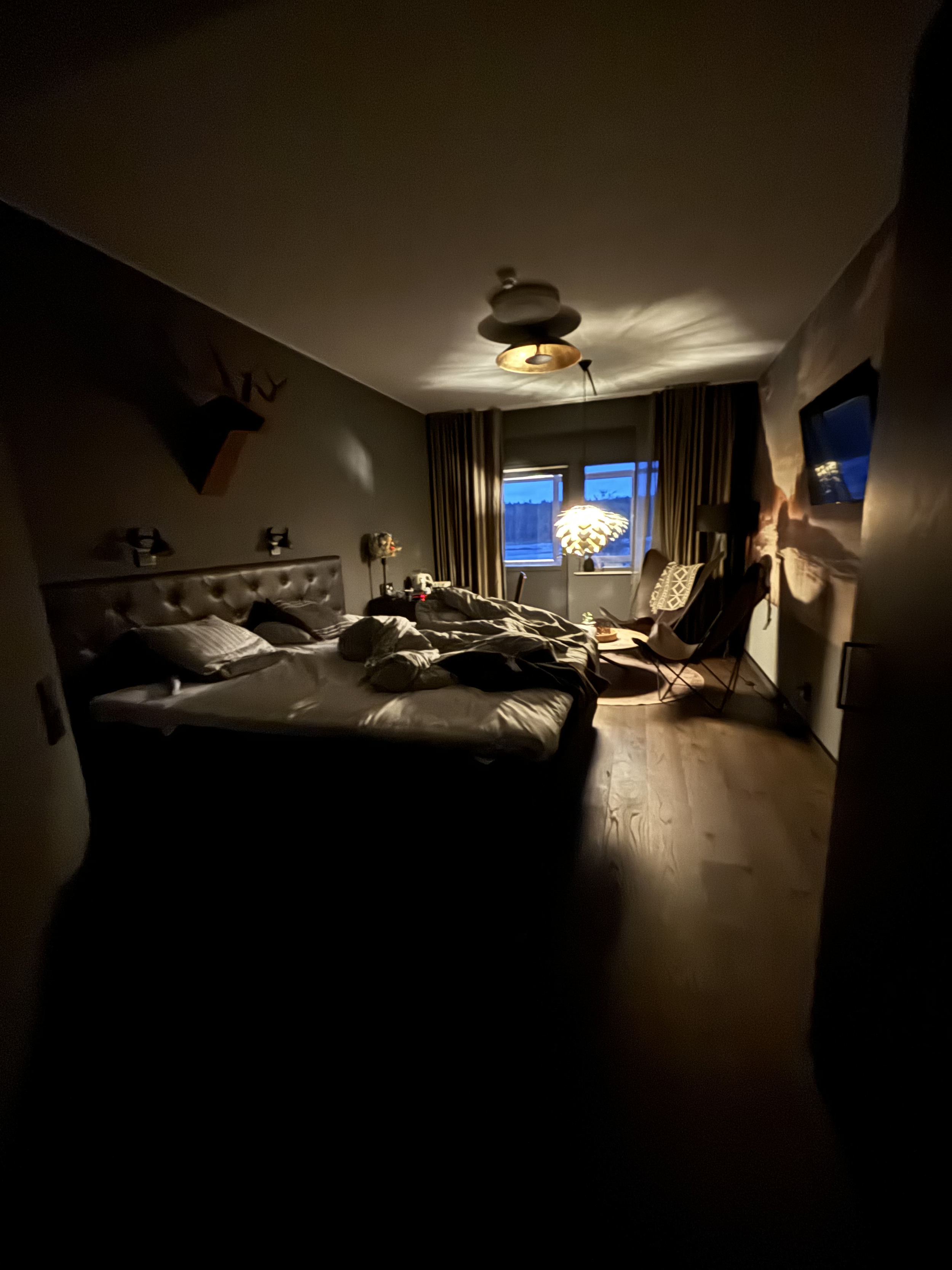 A dimly lit bedroom with a bed, chairs, a window, and a hanging lamp. The bed appears messy with pillows and bedding, and some furniture and objects are visible around the room.