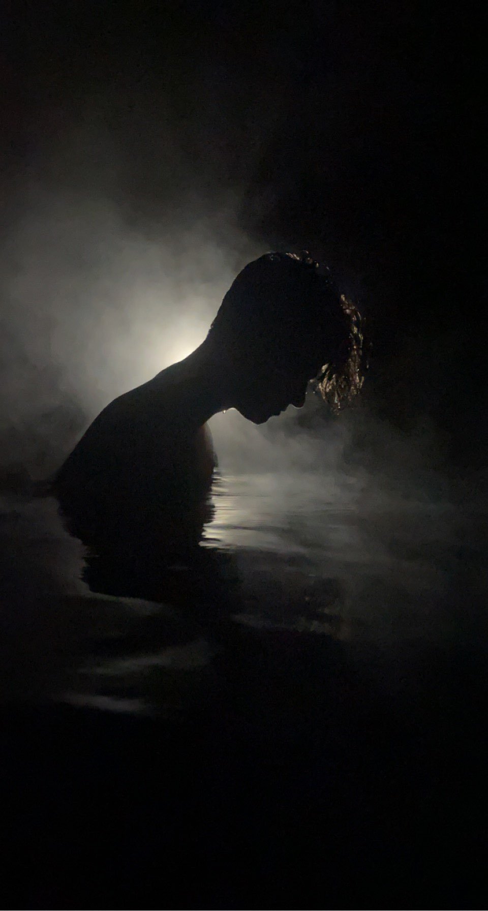 Silhouette of a person with wet hair, head bowed, in water with steam or mist around them, backlit for dramatic effect.
