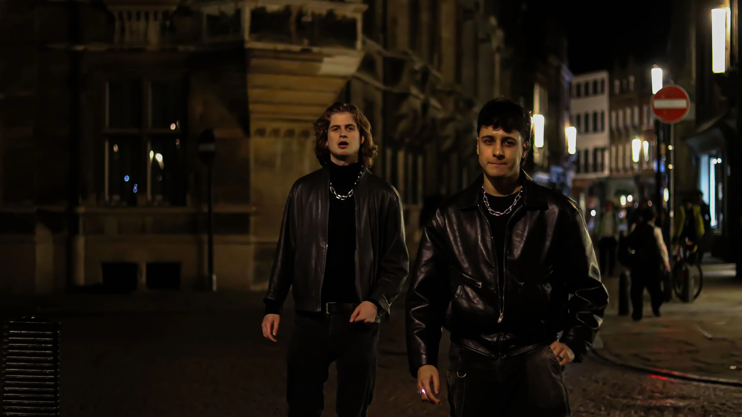 Two young men walking at night on a city street, dressed in black leather jackets with chain necklaces, with buildings and streetlights in the background.