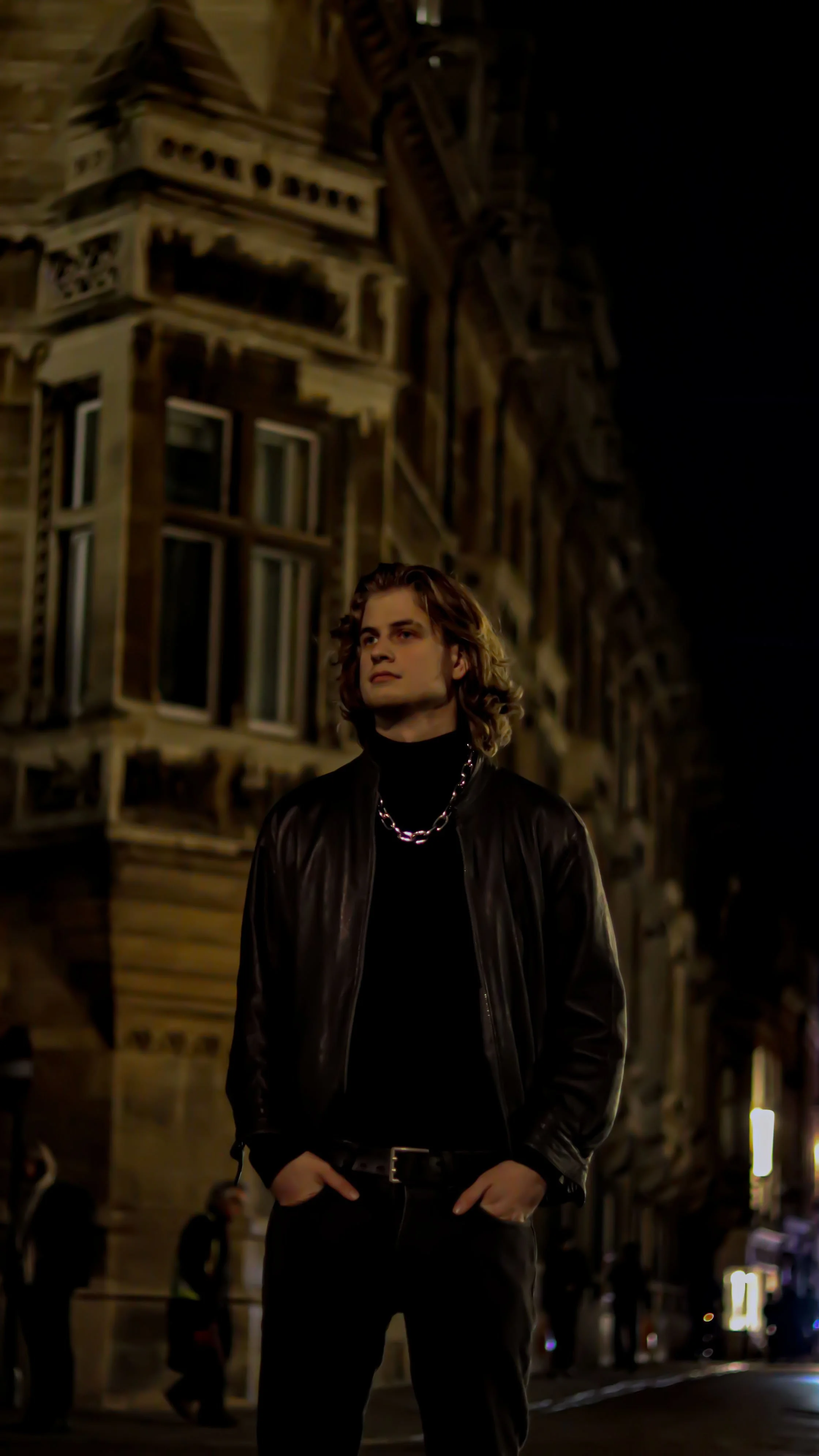 A young man with shoulder-length wavy hair stands on a city street at night, wearing a black leather jacket, black turtleneck, black pants, and a silver chain necklace. There are tall, old buildings with detailed architecture illuminated by streetlig