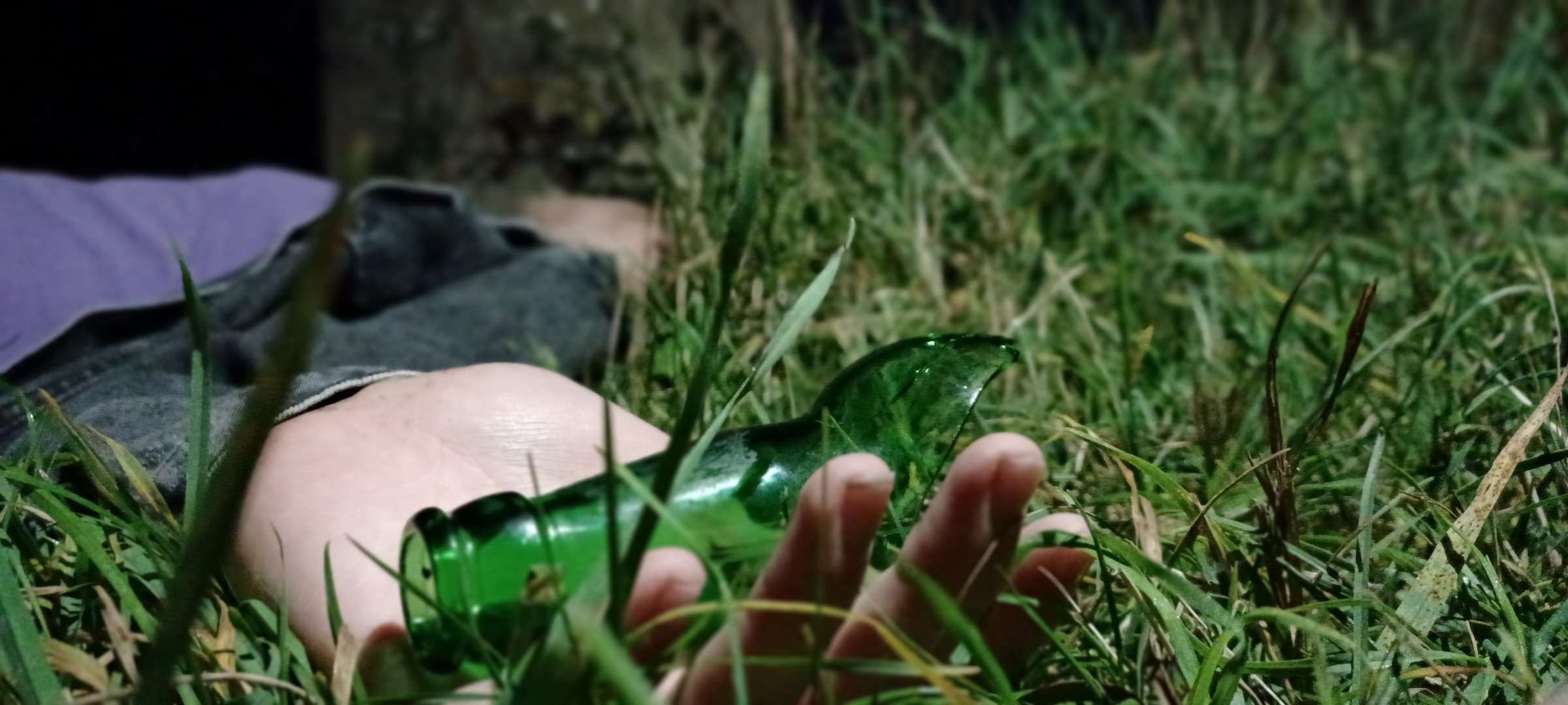Person lying in grass, holding a green glass bottle, with a blurred background of grass and vegetation.