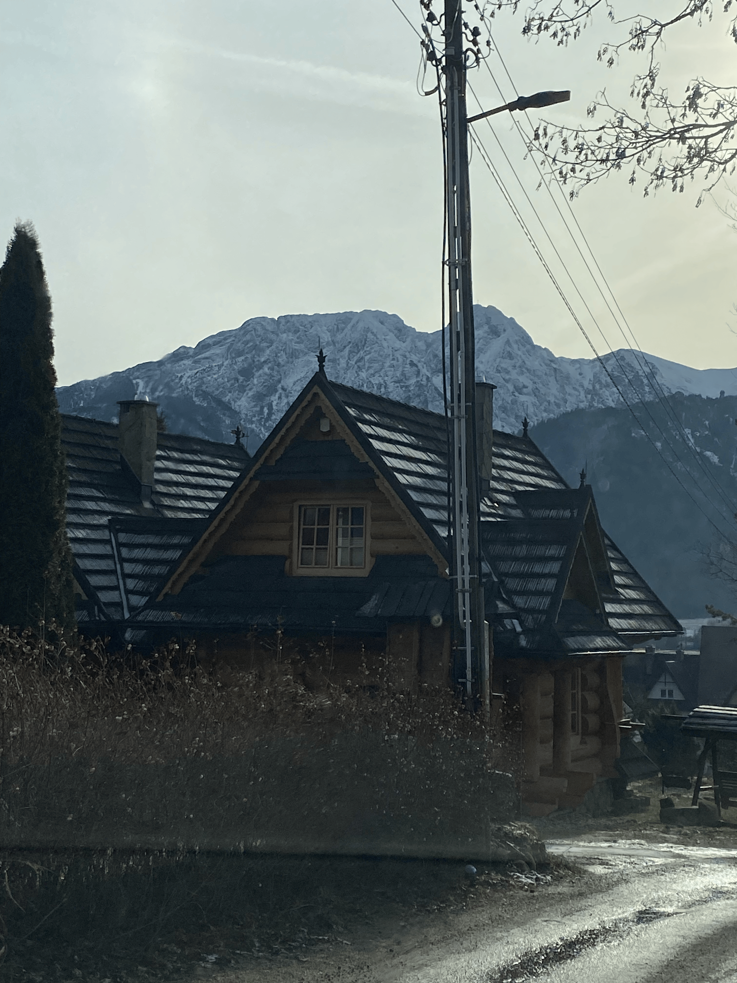 A house with a wooden roof and a window, in front of the house there is a construction site with building materials and a dirt path, with a mountain Stack in the background, electrical power lines and a pole are visible in the foreground.