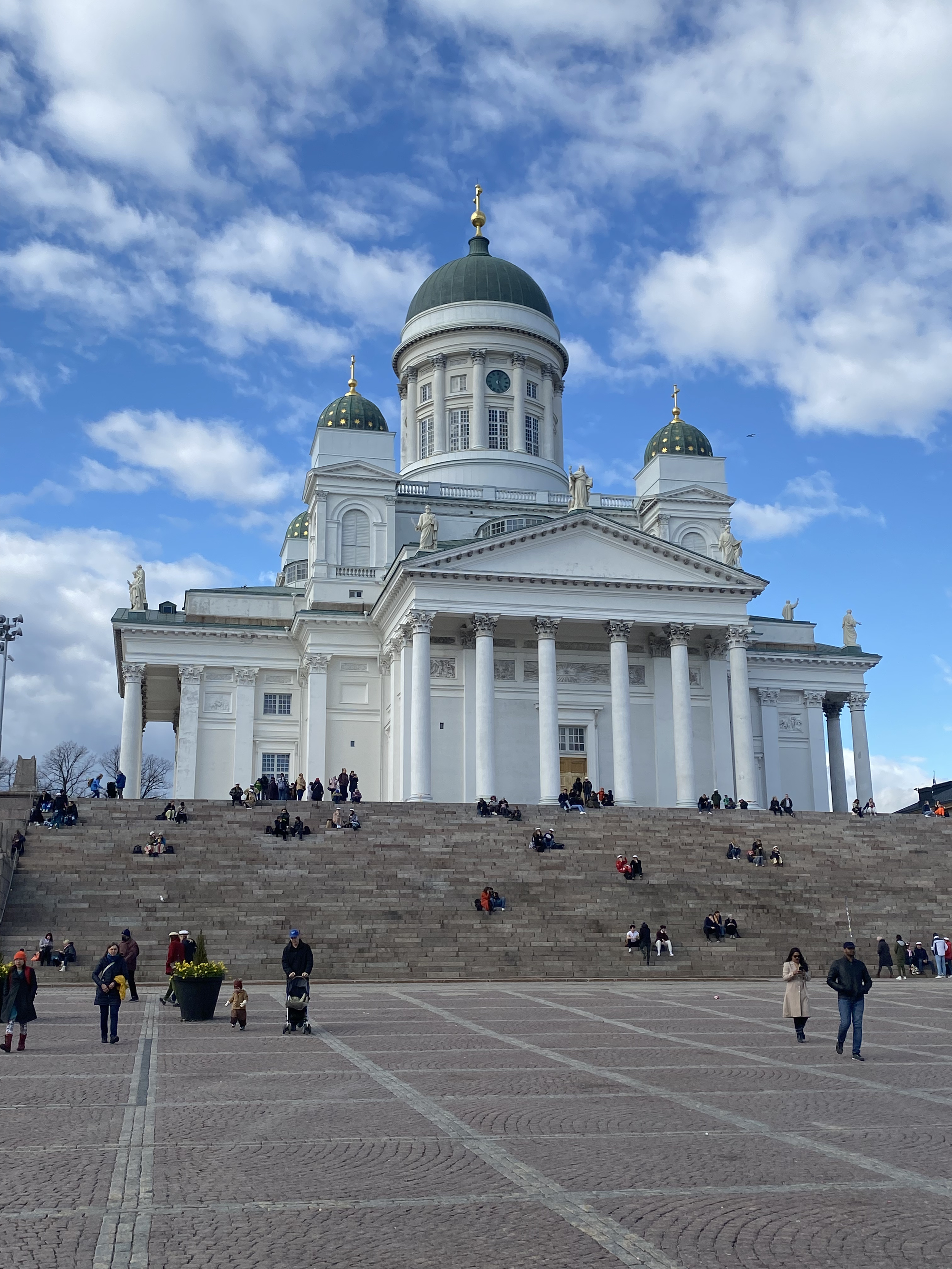 View of a large white church with green domes and gold crosses, with steps leading up to it, and people walking and sitting in the foreground, under a partly cloudy sky.