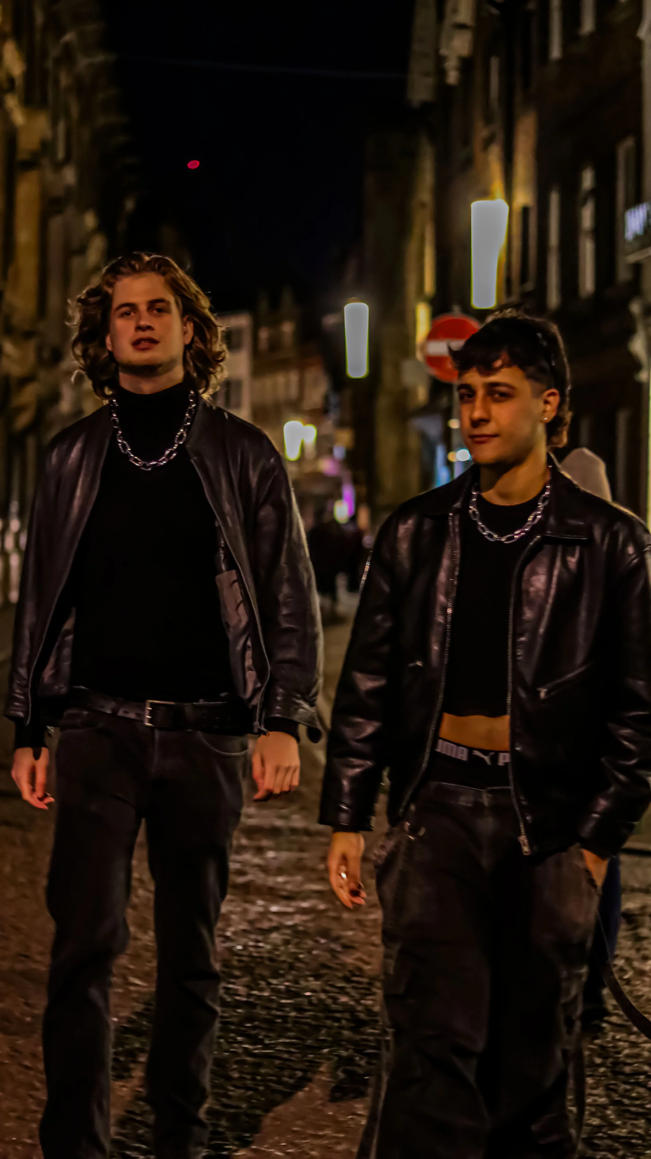 Two young men walking on a city street at night, wearing black leather jackets, black shirts, and chains, with blurred city lights and buildings in the background.