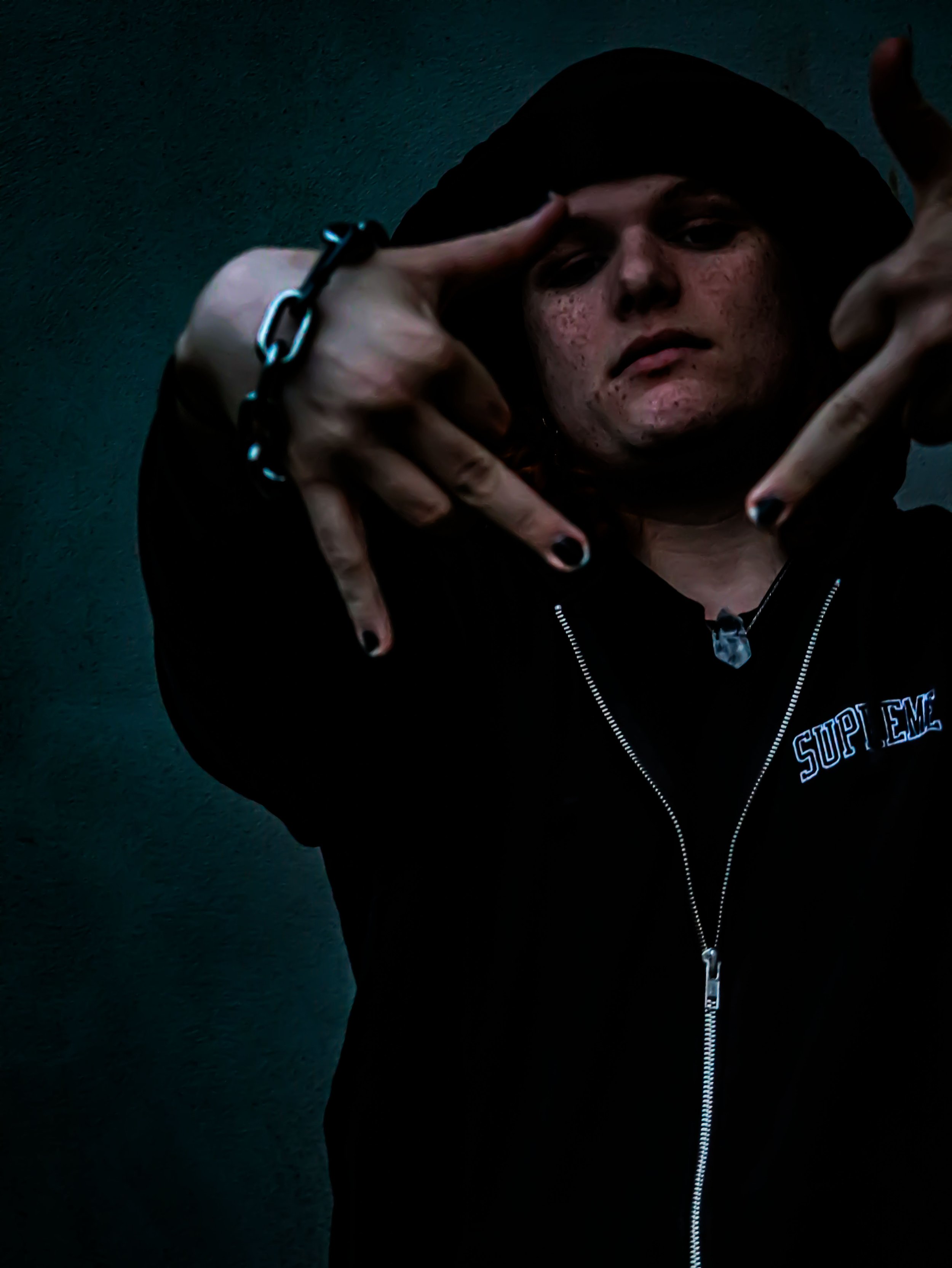 A person wearing a black hoodie with the word 'Supreme' on it, making a hand gesture towards the camera and wearing a chain necklace, standing against a dark background.