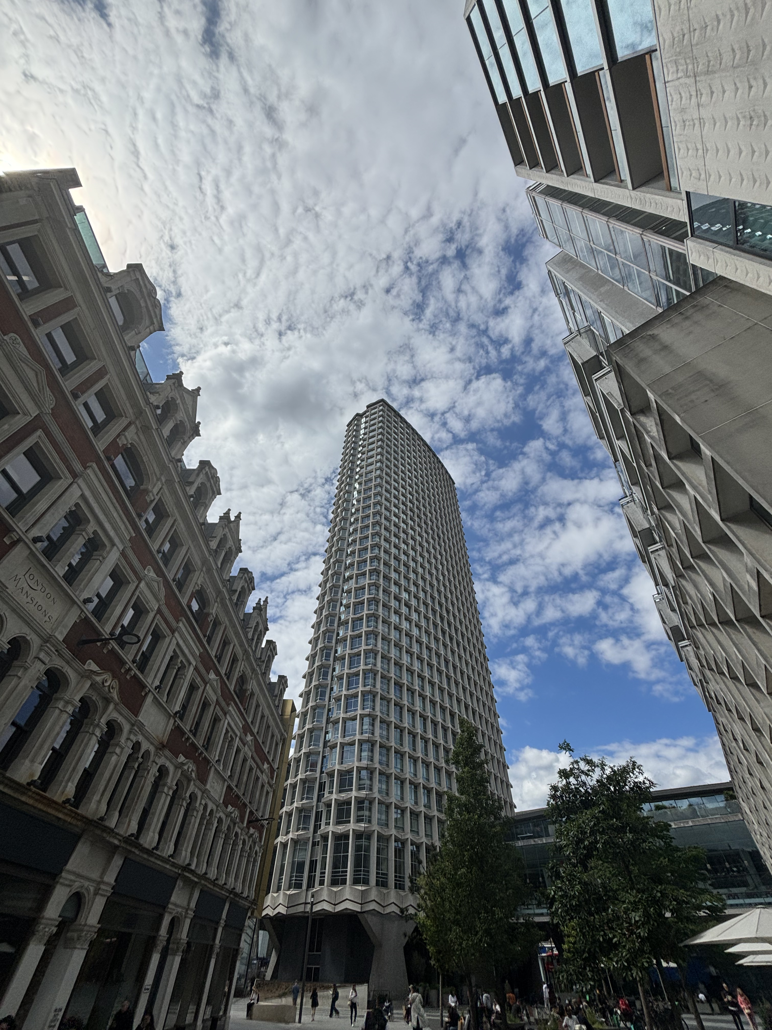 Tall modern skyscraper with hexagonal window pattern in an urban city street, surrounded by historic and contemporary buildings, with pedestrians walking and trees in the foreground under a partly cloudy sky.