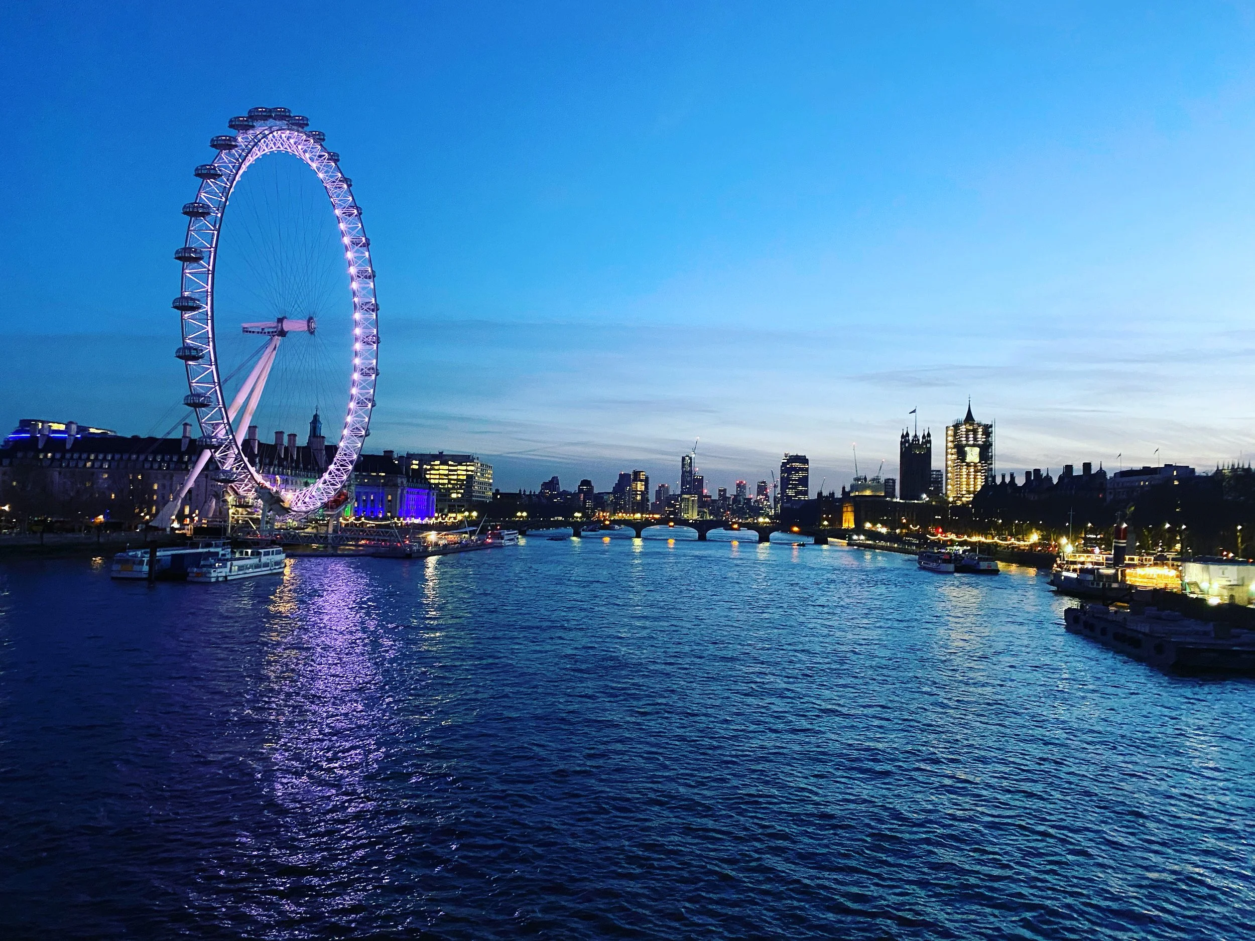 Nighttime view of the London Eye illuminated in purple, with the London skyline and river in the background, including buildings with lights and a clear sky.