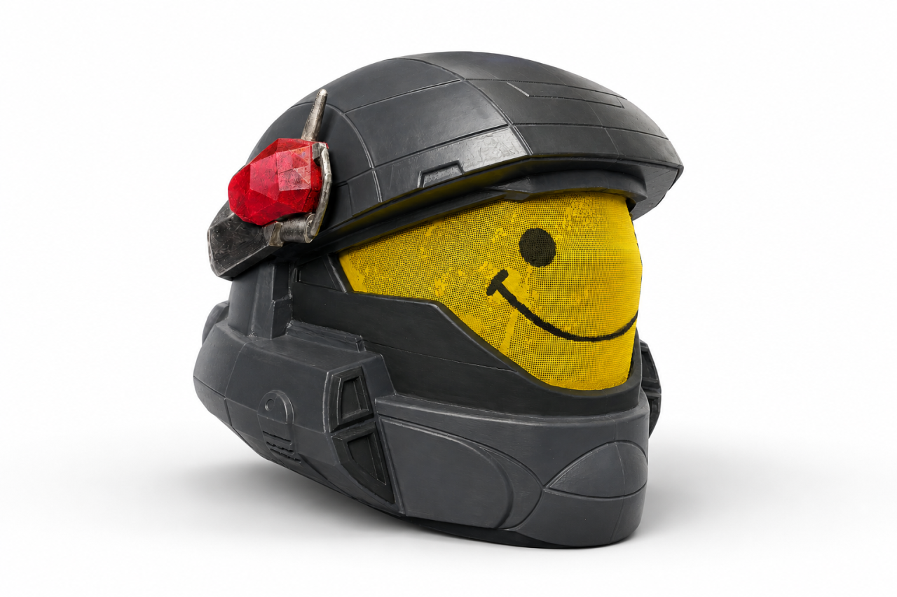 A black realistic helmet with a yellow smiley face visor and a red gem on the side.