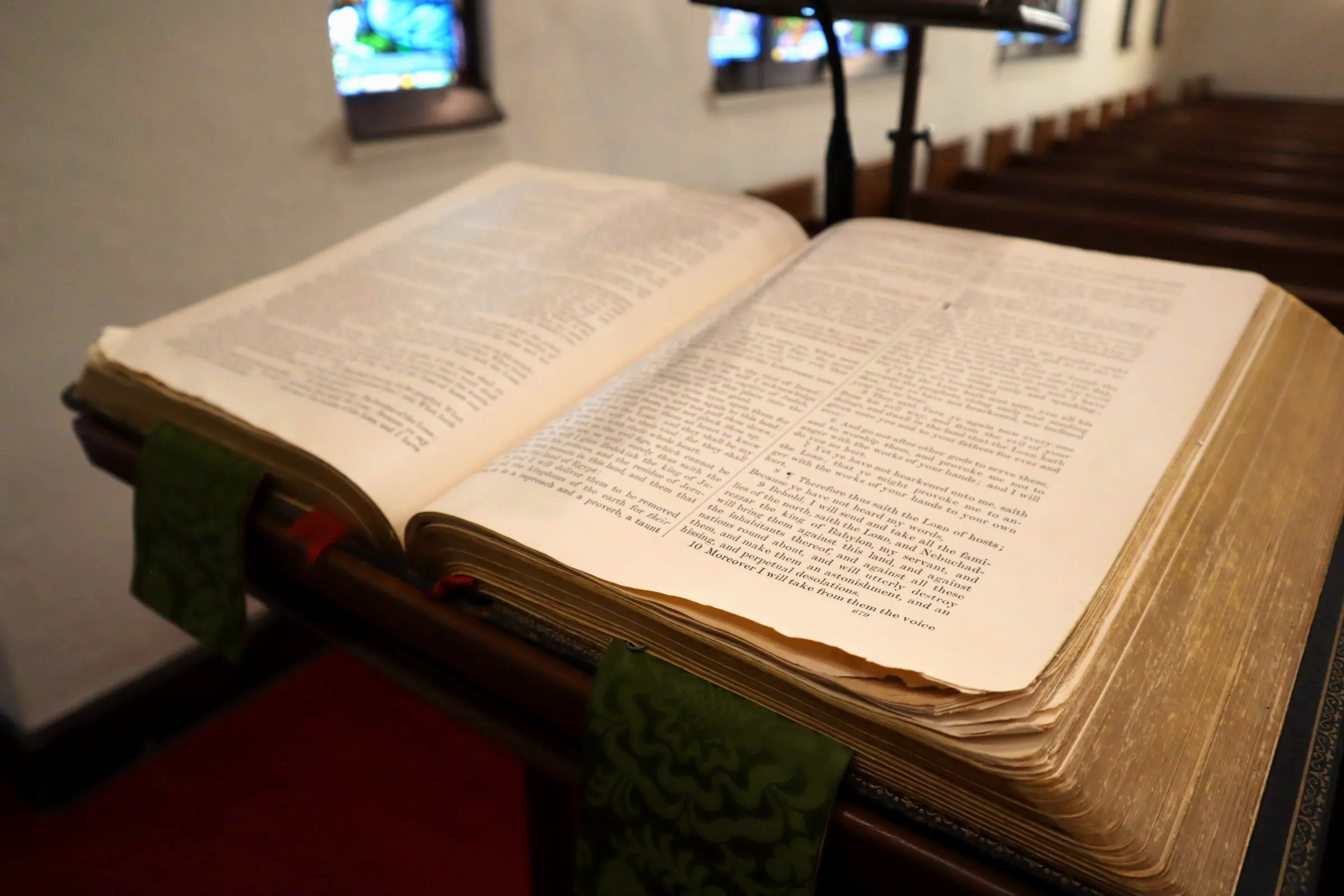 An open Bible resting on a wooden lectern in a church or chapel setting, with stained glass windows visible in the background.