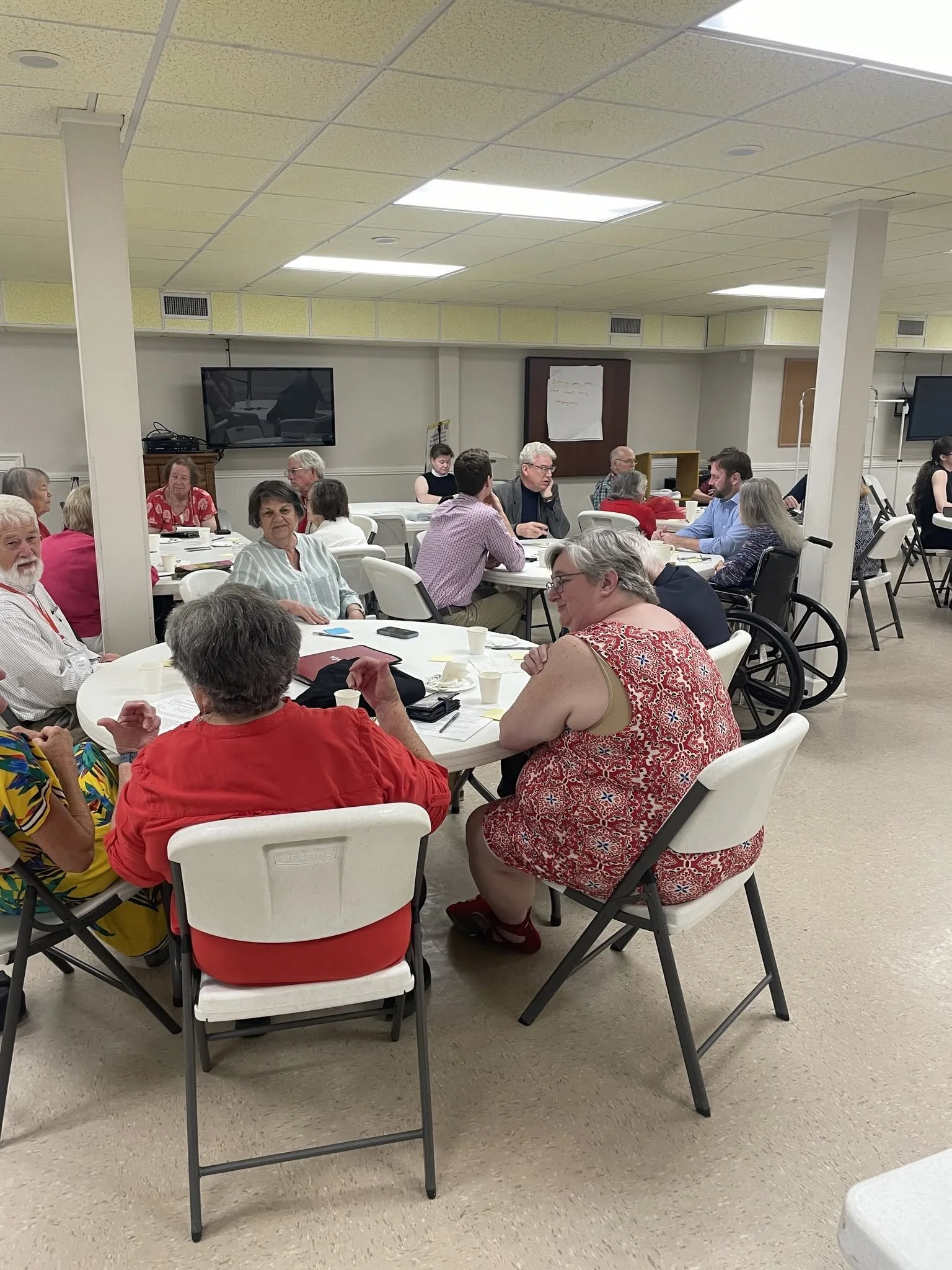 A group of people sitting around tables in a conference room, engaging in discussion or listening to a speaker, with some individuals in wheelchairs and a large TV screen on the wall in the background.