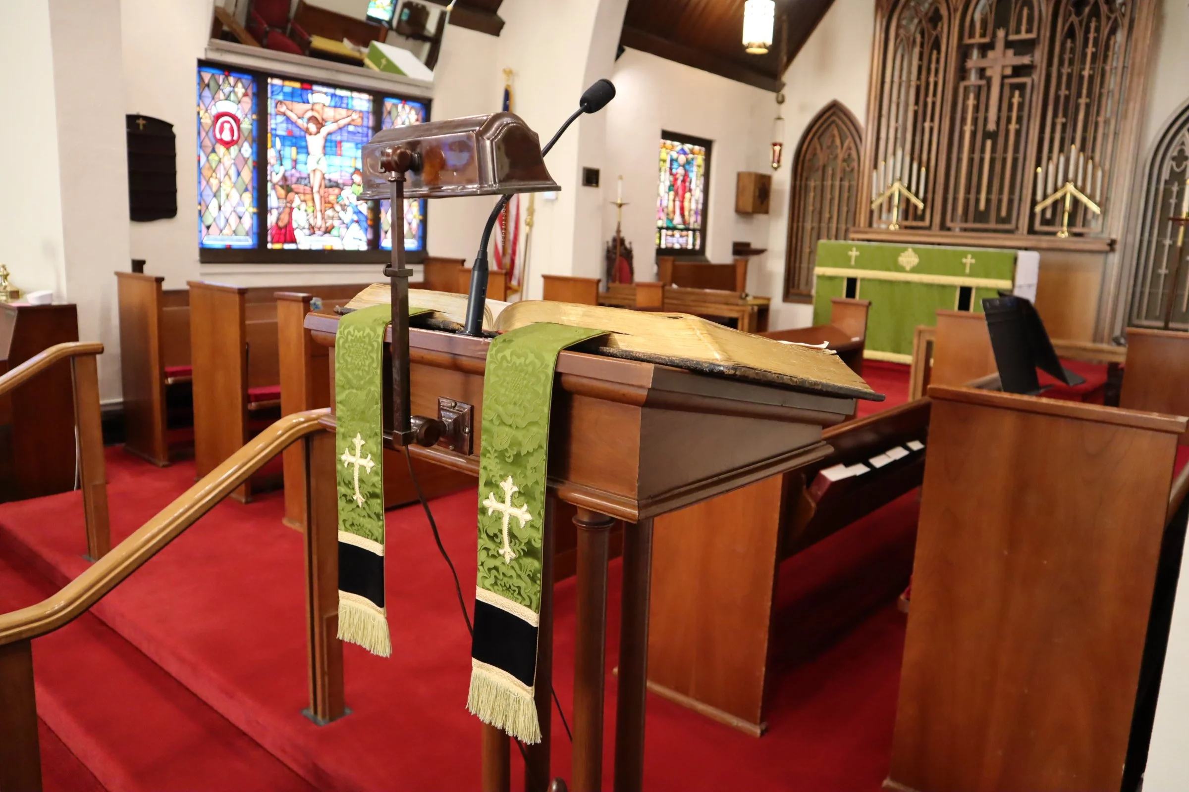 Inside a church, a wooden pulpit with a green liturgical cloth with white crosses draped over it. In the background, stained glass windows depicting religious scenes, wooden pews with red cushions, and a decorative wooden screen with gold accents. A microphone is attached to the pulpit.