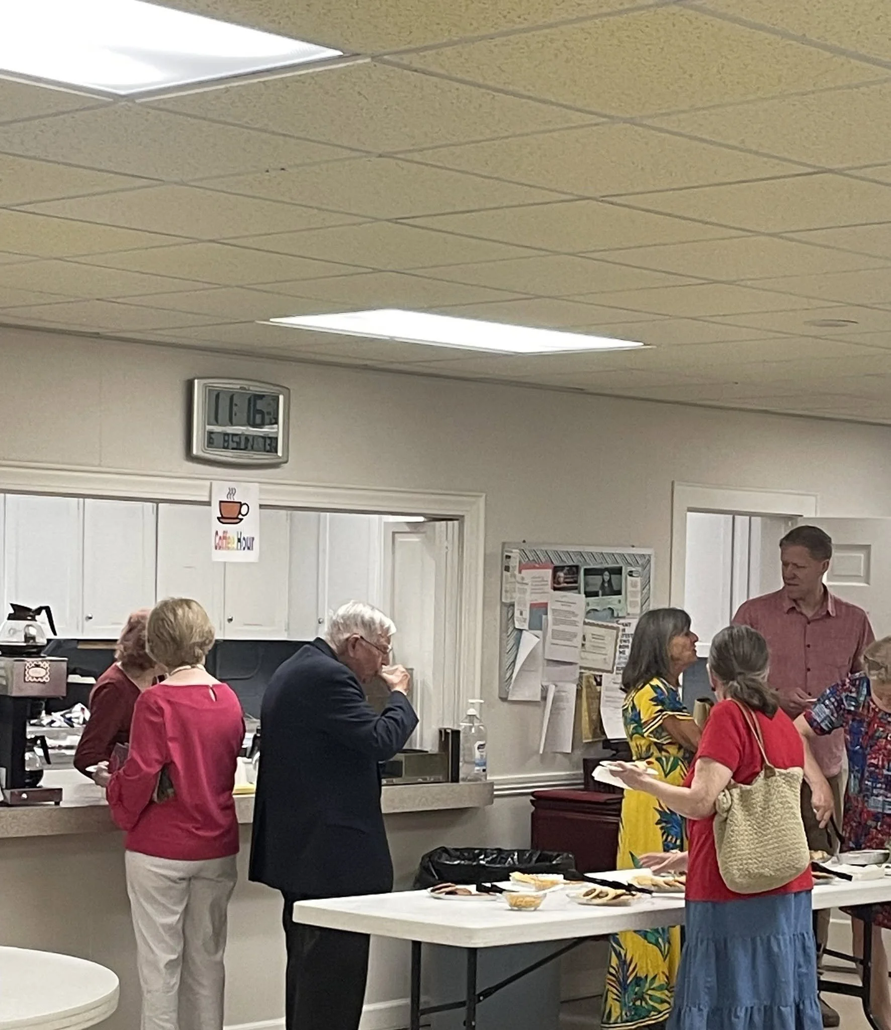 People gathered around a serving table with food, some serving, some engaging in conversation, in an indoor setting with ceiling lights and a wall-mounted clock.